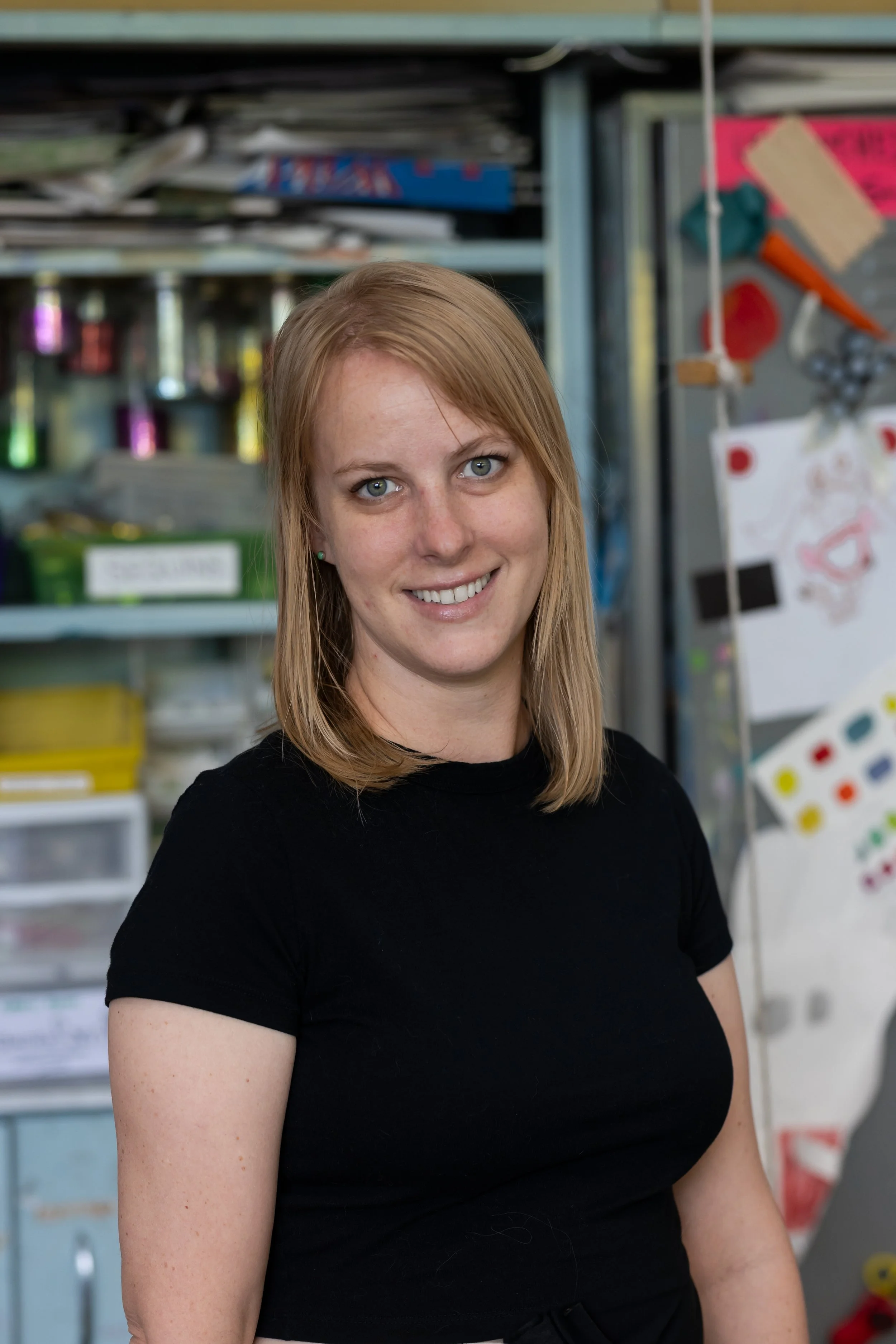 A woman with blonde hair and green eyes wearing a black shirt, smiling, standing in what appears to be a classroom or art space with shelves and colorful art supplies in the background.