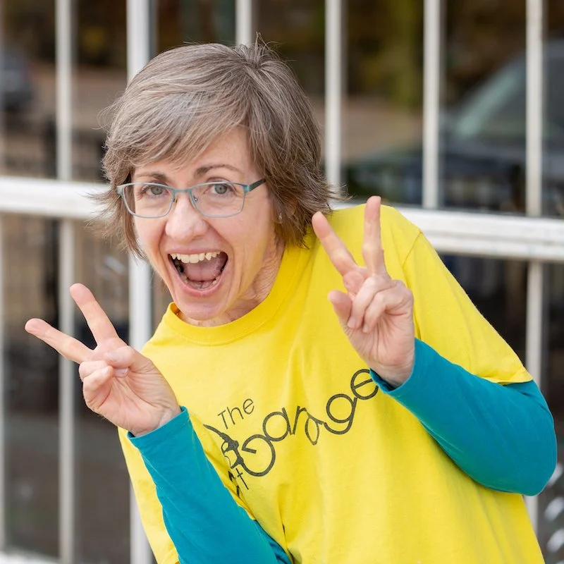 Woman with short gray hair, glasses, wearing a yellow T-shirt and blue long-sleeve shirt underneath, making peace signs with both hands and smiling.