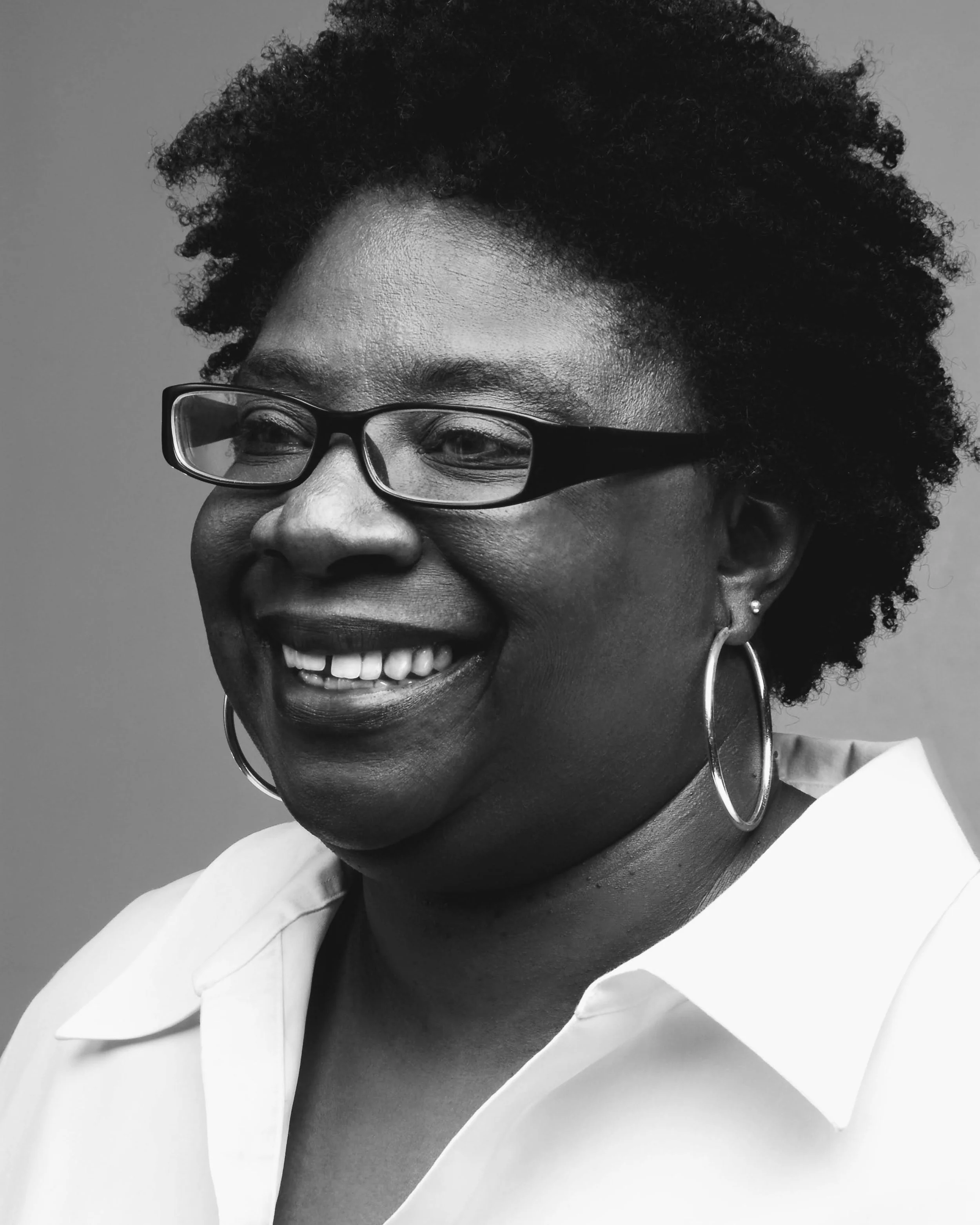 Close-up of a Black woman smiling, wearing glasses, hoop earrings, a white shirt, and a dark top, with short, curly hair.
