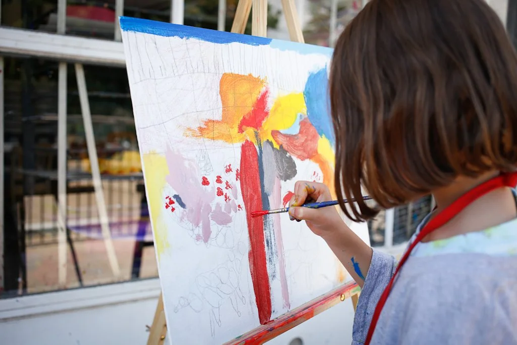 A young girl painting on a canvas outdoors, with various colors including red, orange, yellow, and blue, on a cloudy day.
