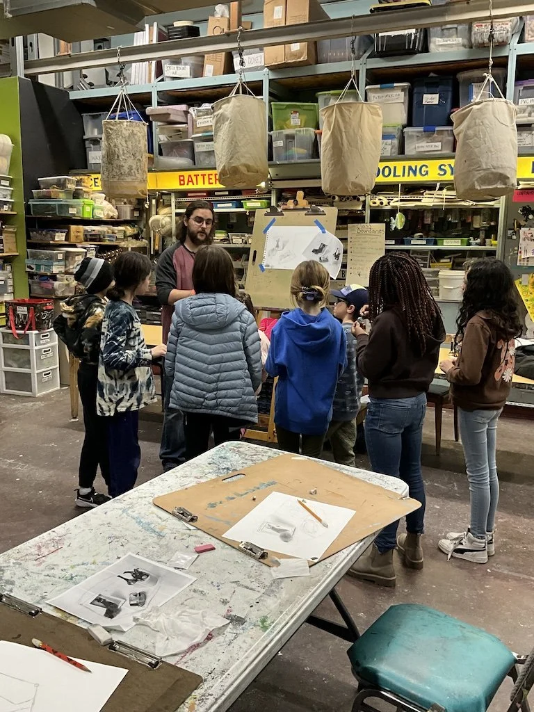 A group of children gathered around a man in a workshop or classroom, with shelves containing supplies and hanging paper bags in the background.