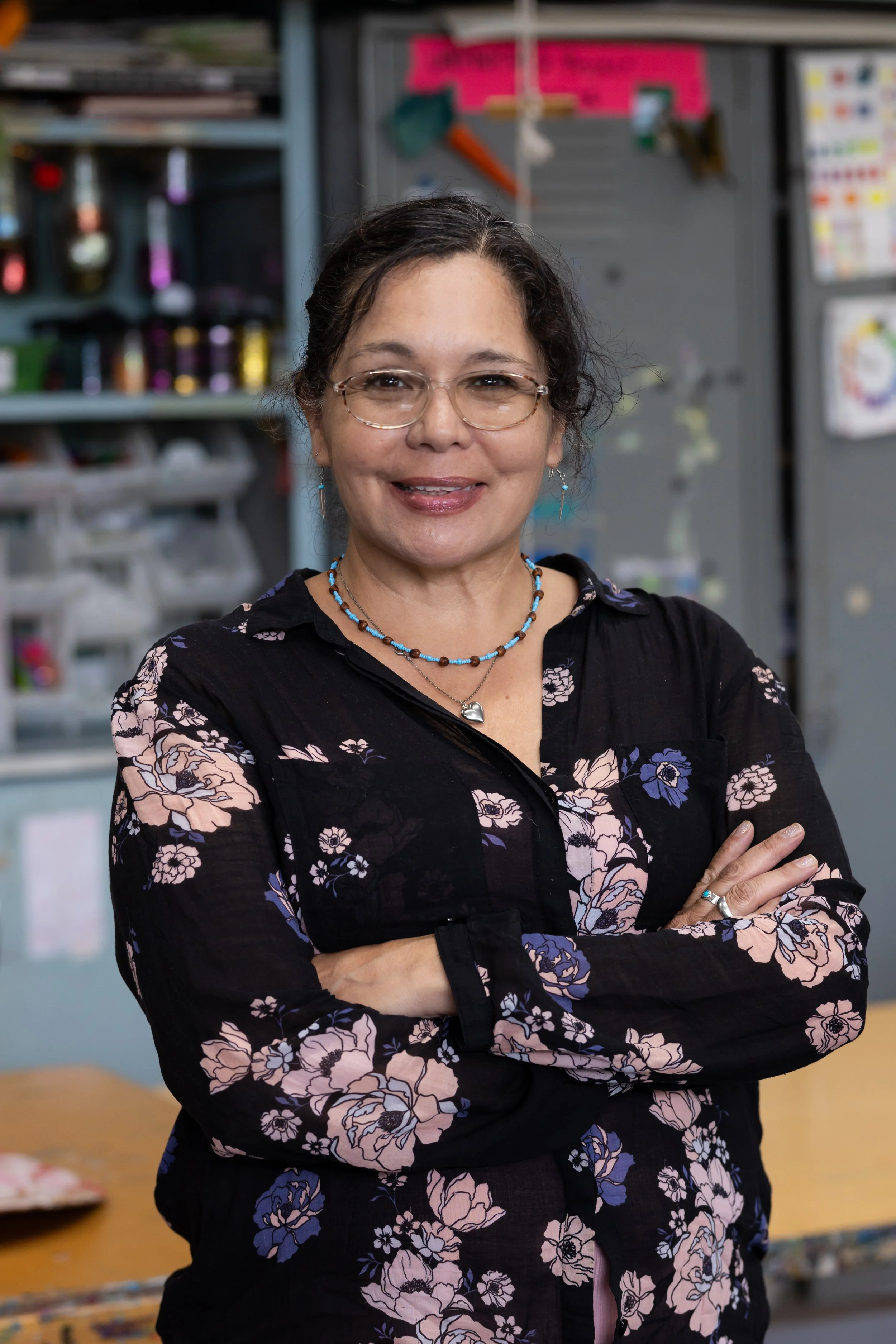 A woman with glasses, wearing a black floral shirt and jewelry, standing with arms crossed in an art or craft room with shelves of supplies in the background.