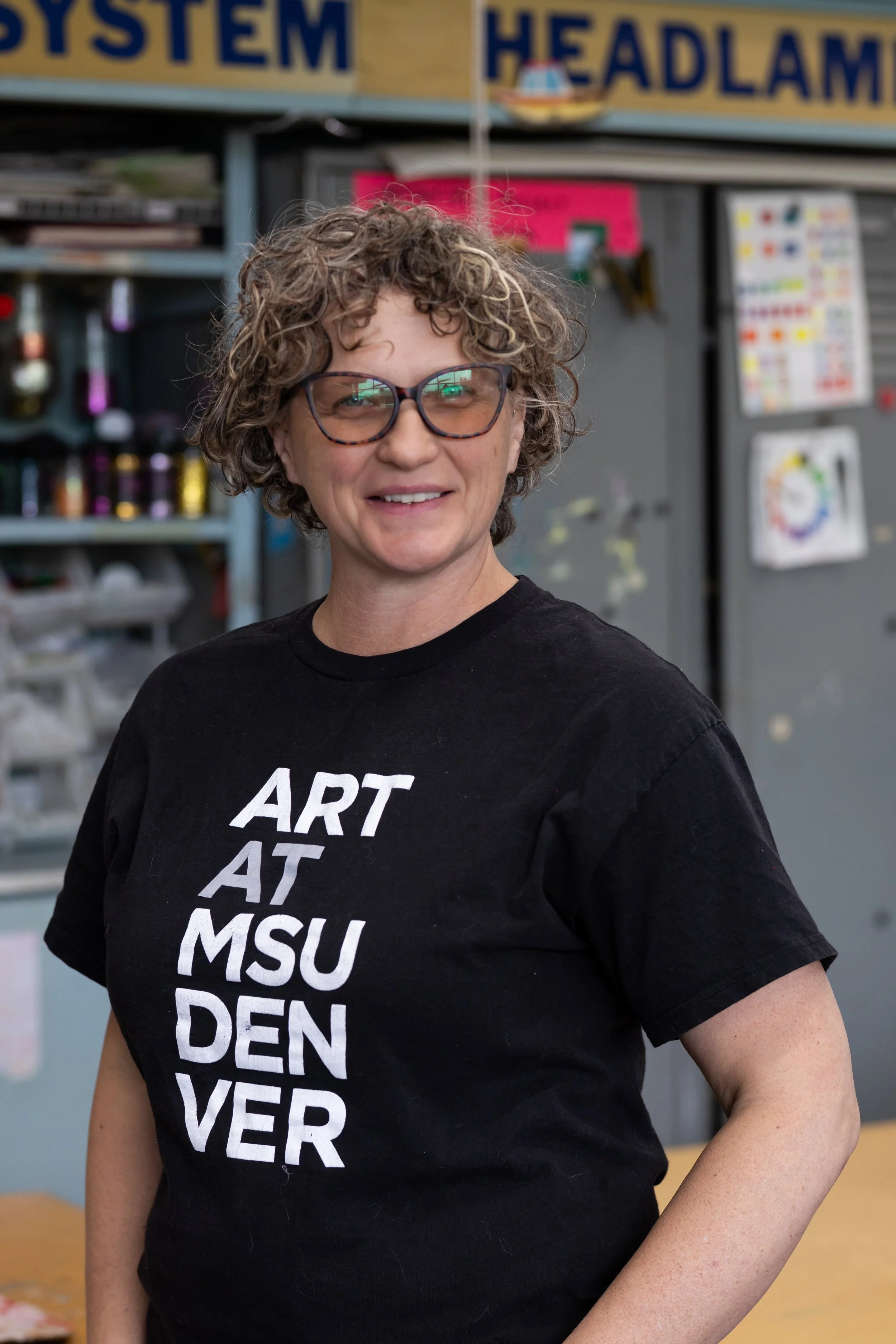 A woman with short curly hair and glasses smiling in front of a display with art supplies and signs, wearing a black t-shirt with white text.