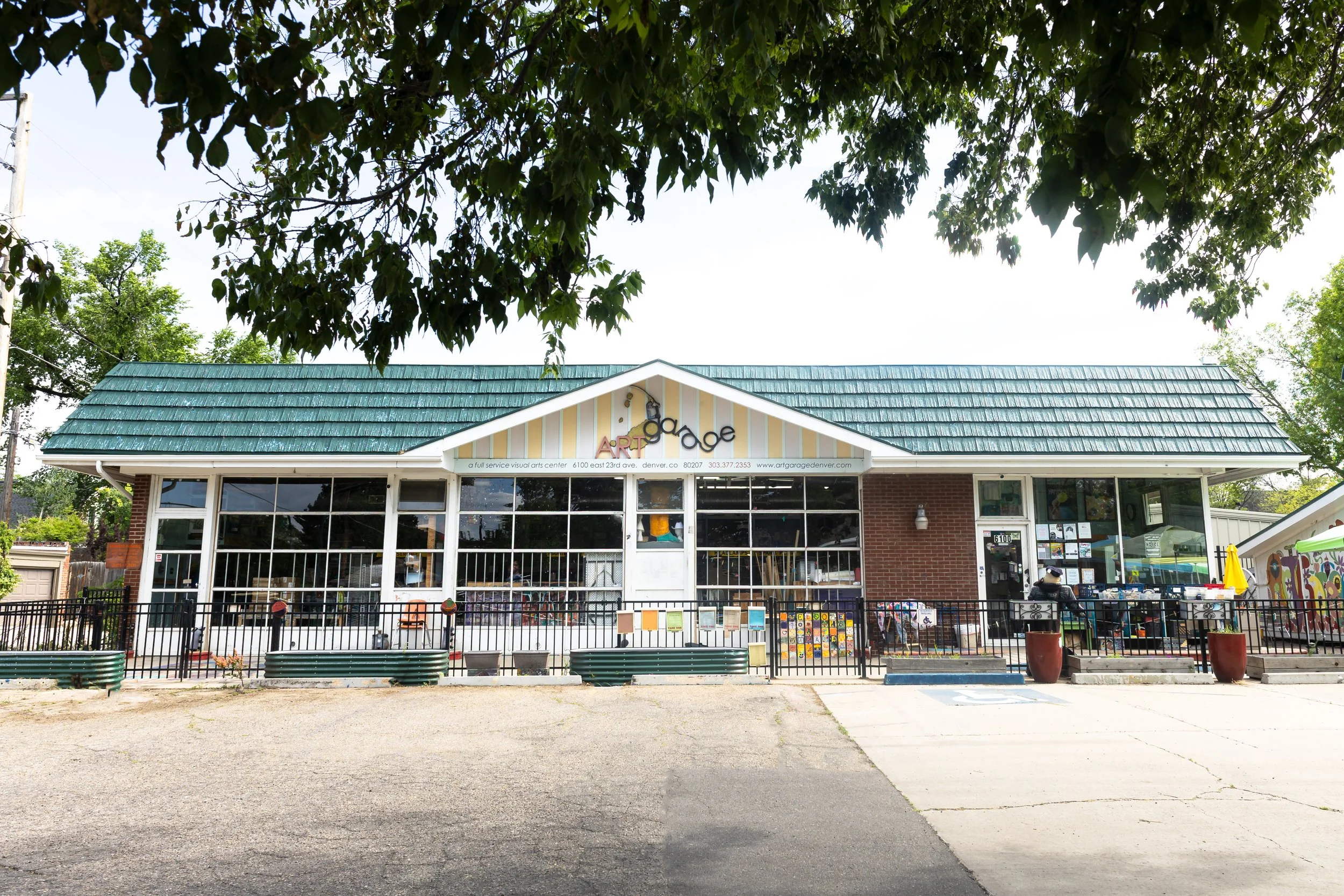 An art gallery building with a teal shingle roof, a large front window, and a sign that reads 'art garage'. There is a black fence in front of the building, with potted plants and artwork displayed along it. The sky is partly cloudy with trees overhead.