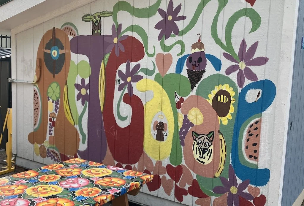 Colorful mural featuring flowers, a tiger, a skull with a cherry on top, a bee, and other whimsical elements painted on a wall.