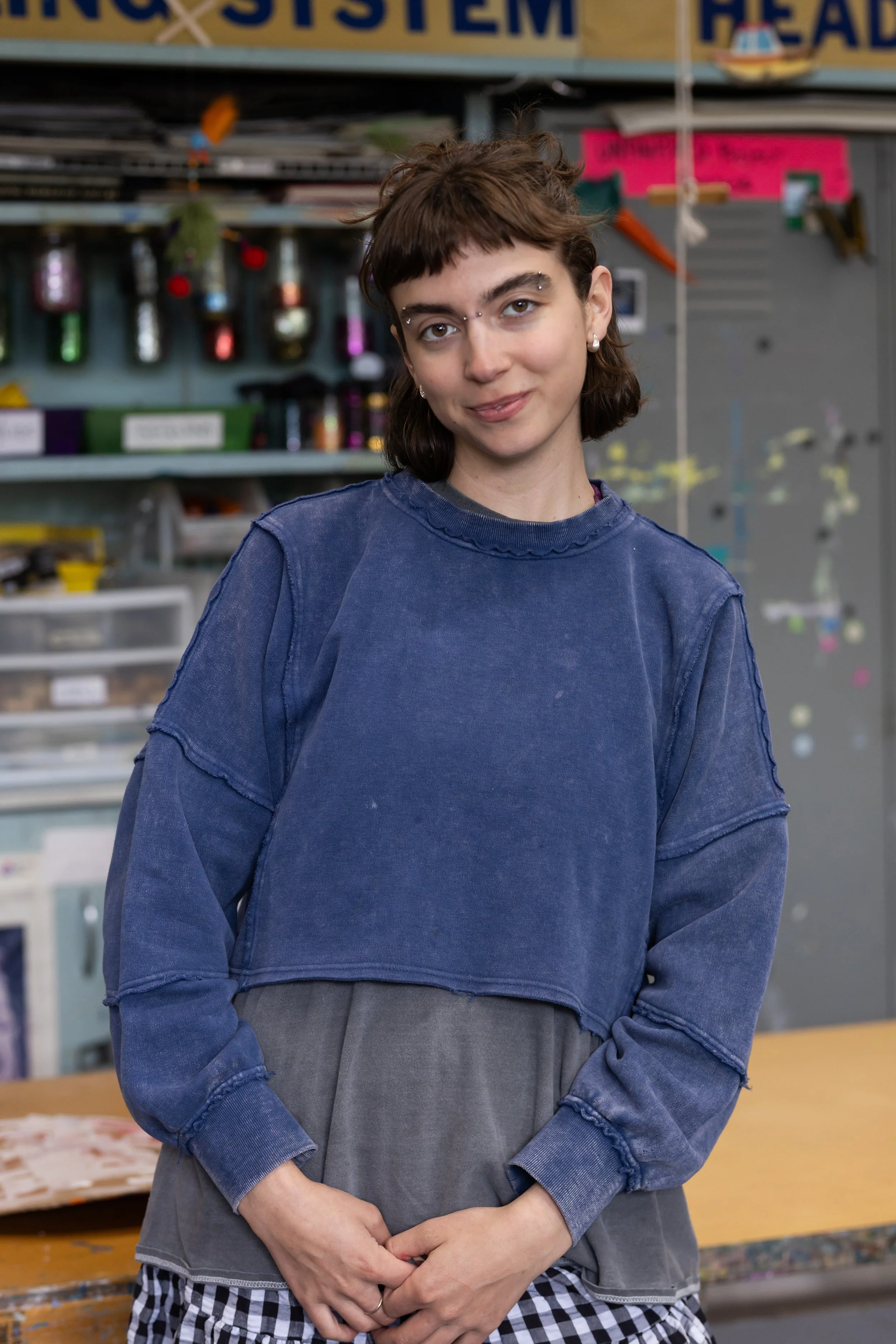 A woman with short, dark hair and a nose piercing, wearing a blue sweatshirt and checkered pants, standing in a workshop with tools and colorful bottles on shelves in the background.