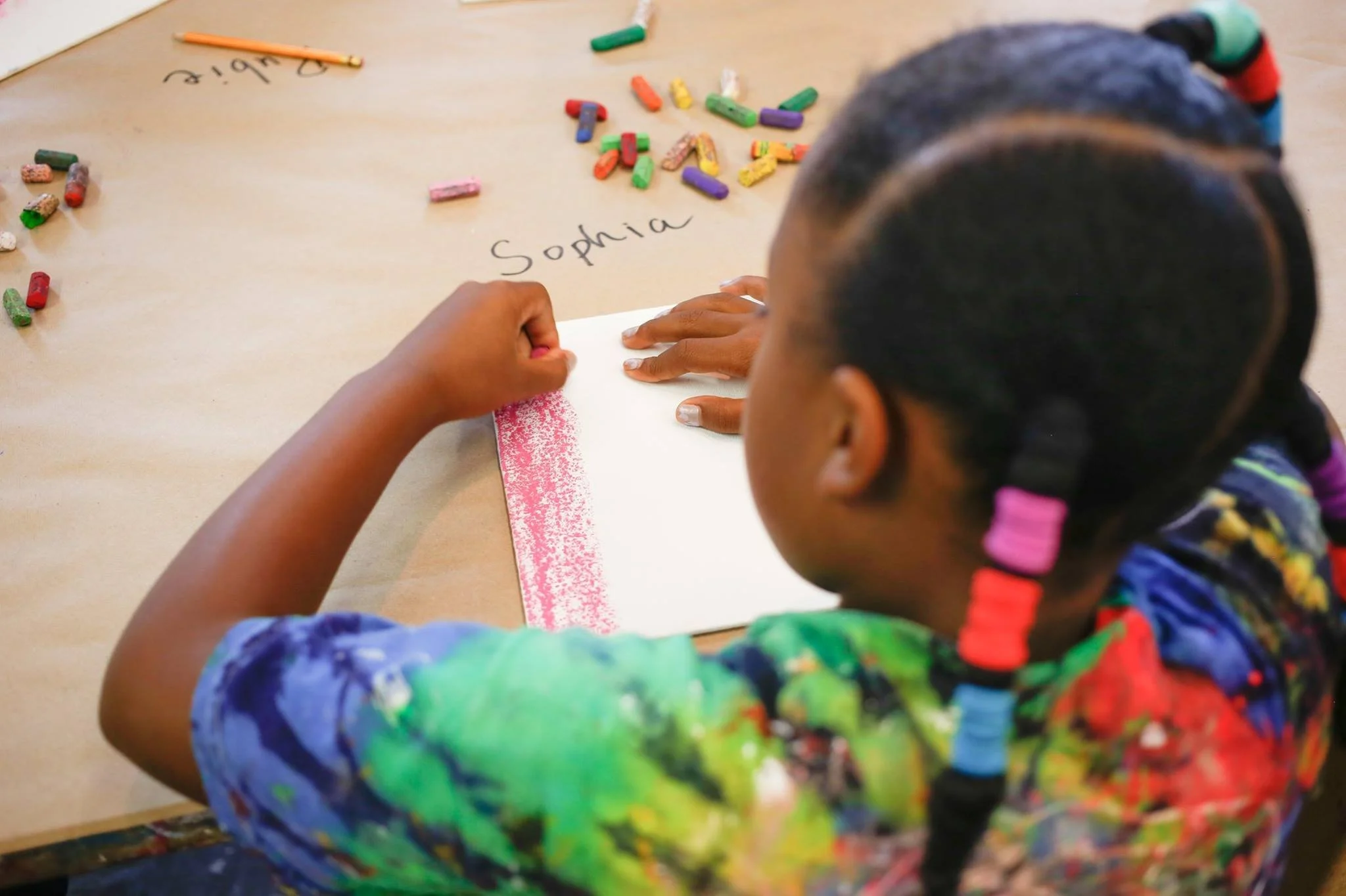 Child with colorful braided hair writing in a notebook surrounded by crayon shavings, with the name Sophia written on the paper.