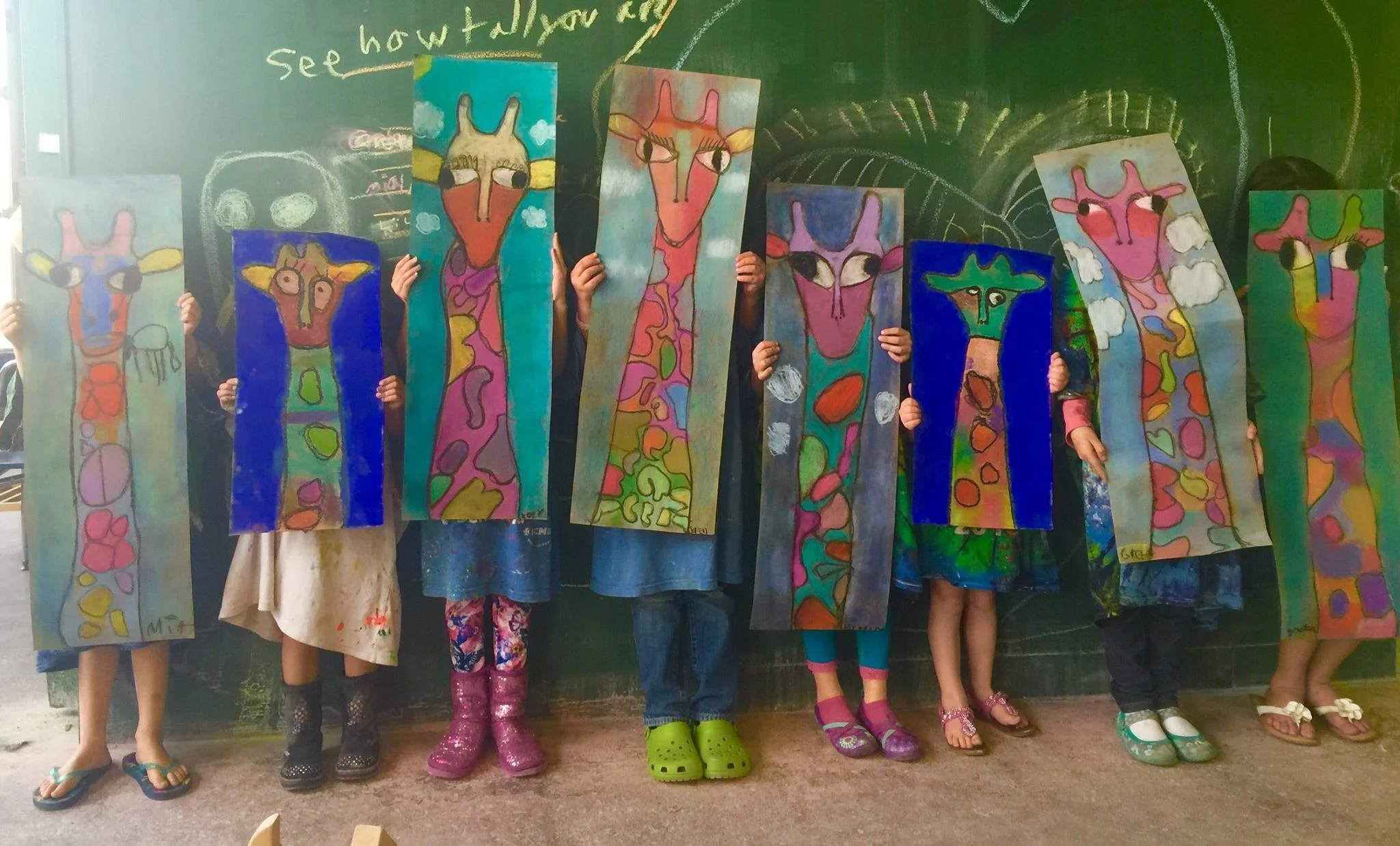 Children standing in a classroom holding up colorful paintings of giraffes with a blackboard in the background.