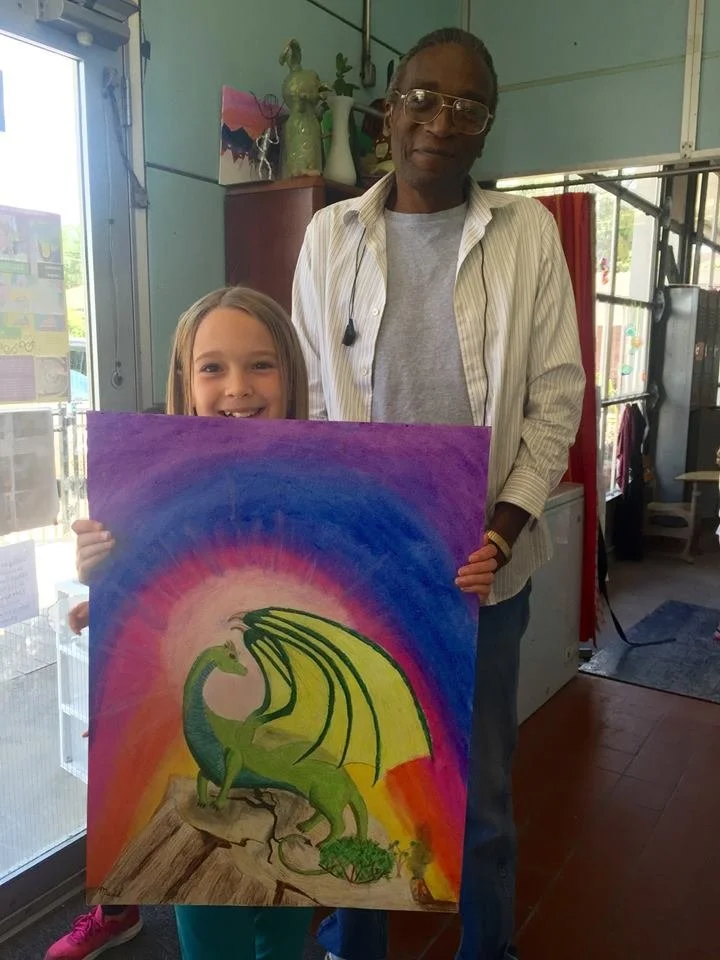A young girl and an older woman holding a colorful painting of a green dragon with yellow wings, standing inside a room with art supplies and decorations.