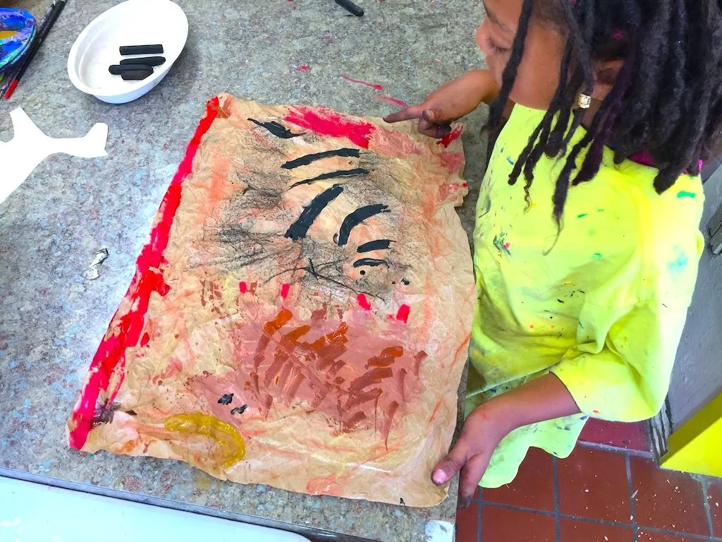 Child in a yellow smock painting a portrait on crumpled brown paper with black and orange paint, using a finger or brush, at a table with art supplies.
