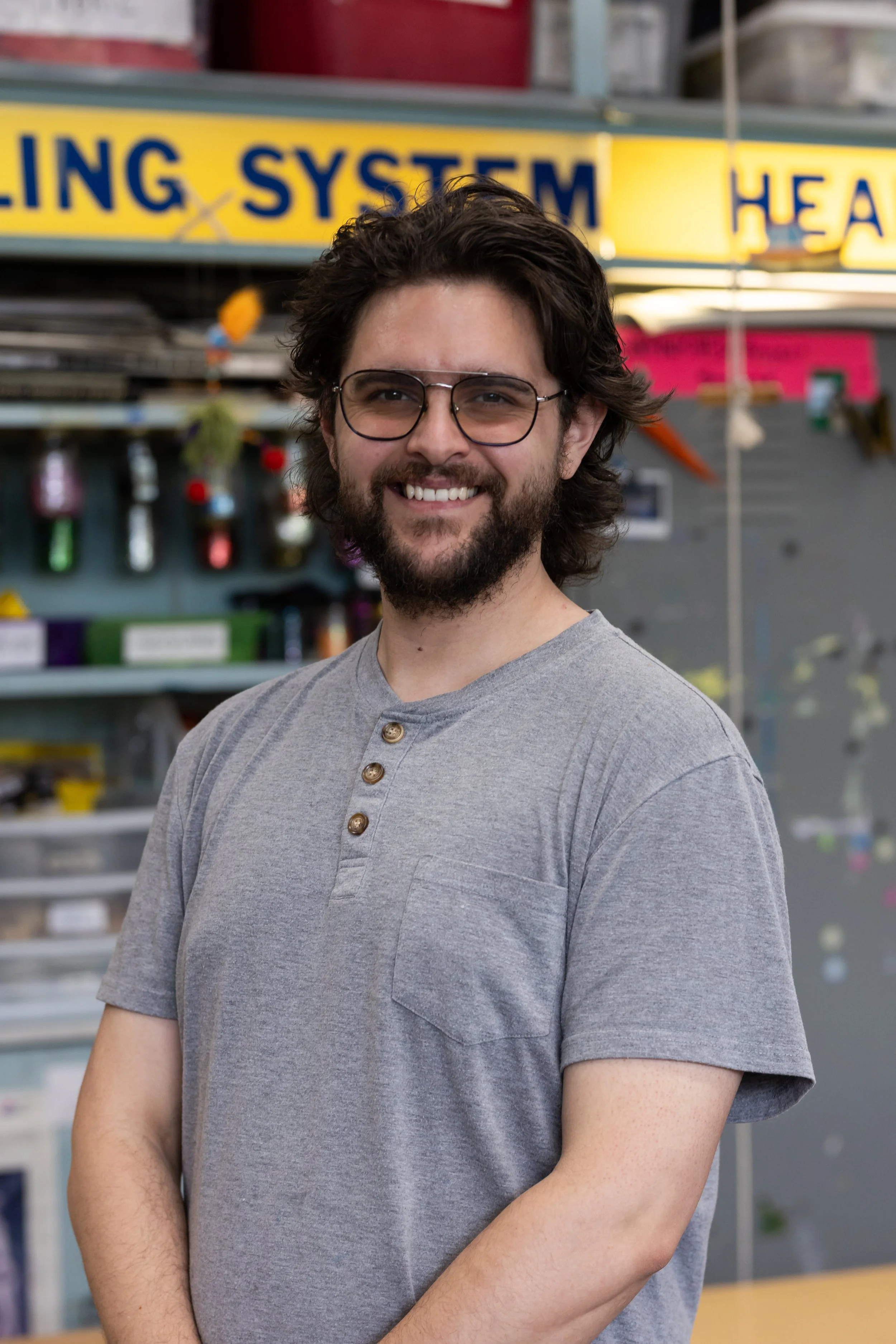 A man with dark, wavy hair, glasses, and a beard smiling, wearing a gray shirt with buttons near the neckline, standing in front of a store display with various tools and equipment.