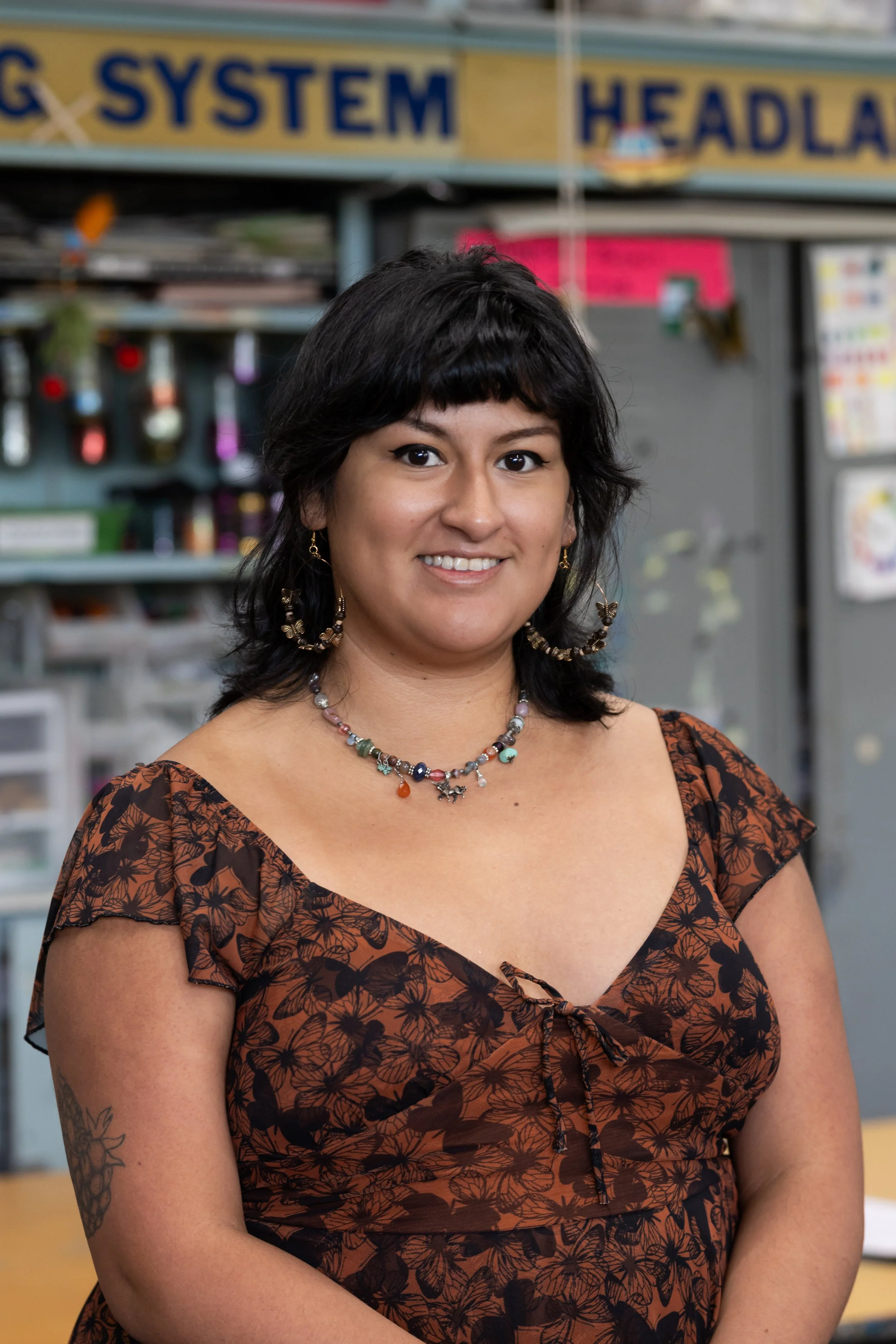 A woman with dark hair and earrings, wearing a butterfly-patterned dress, standing in front of a display with signs and shelves in an art studio.