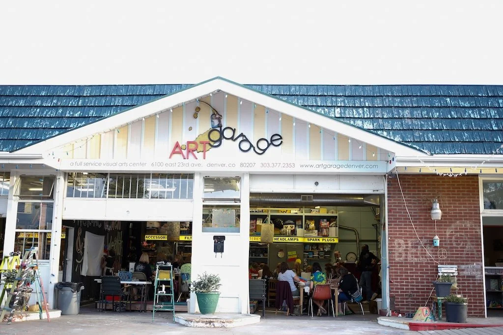 The front view of Art Garage, a visual arts center in Denver, Colorado, with an open garage door showing people inside, art supplies, and display shelves.