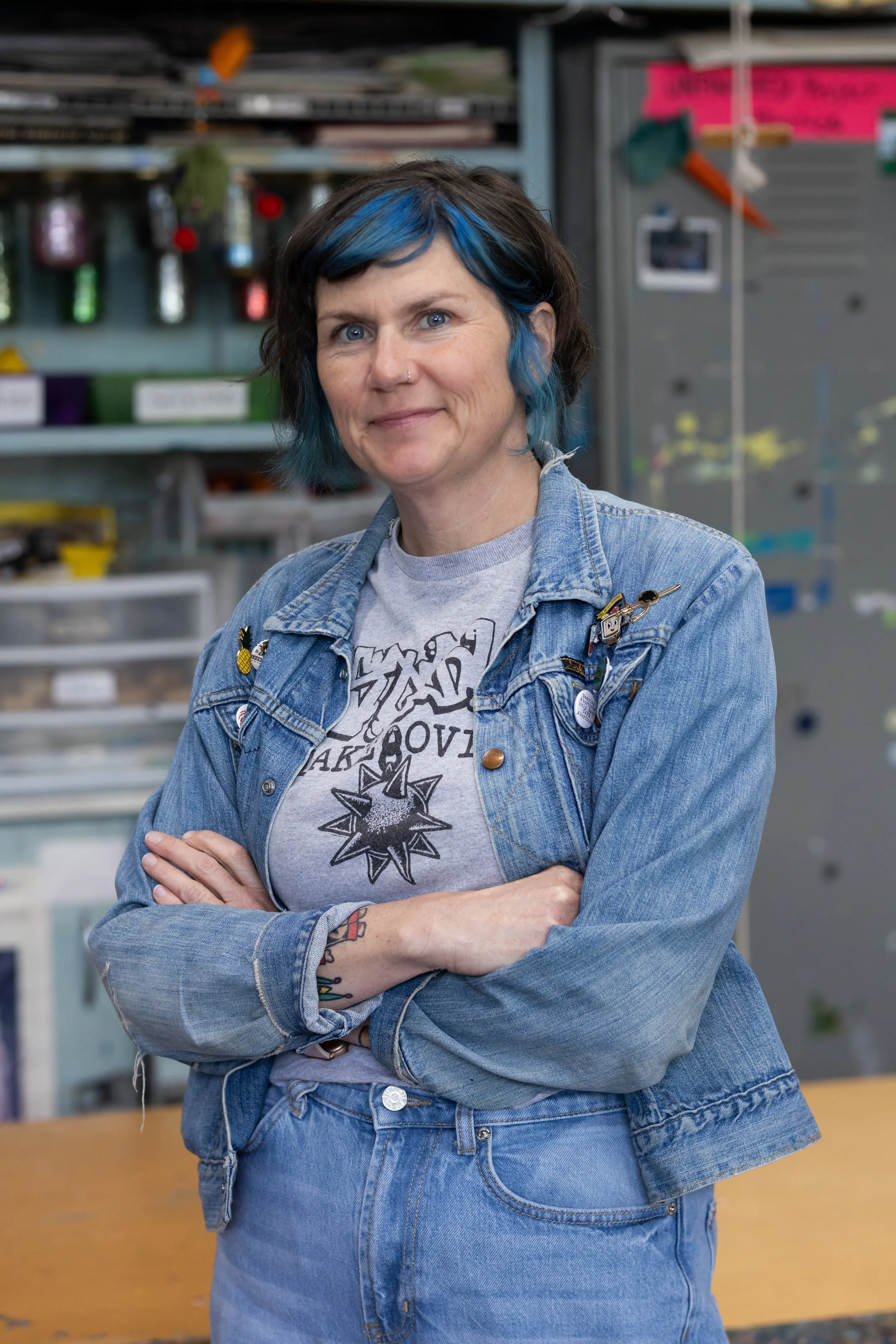 Person with short dark hair and blue streaks, wearing a denim jacket with pins, crossing arms, standing in a workshop or hardware store.