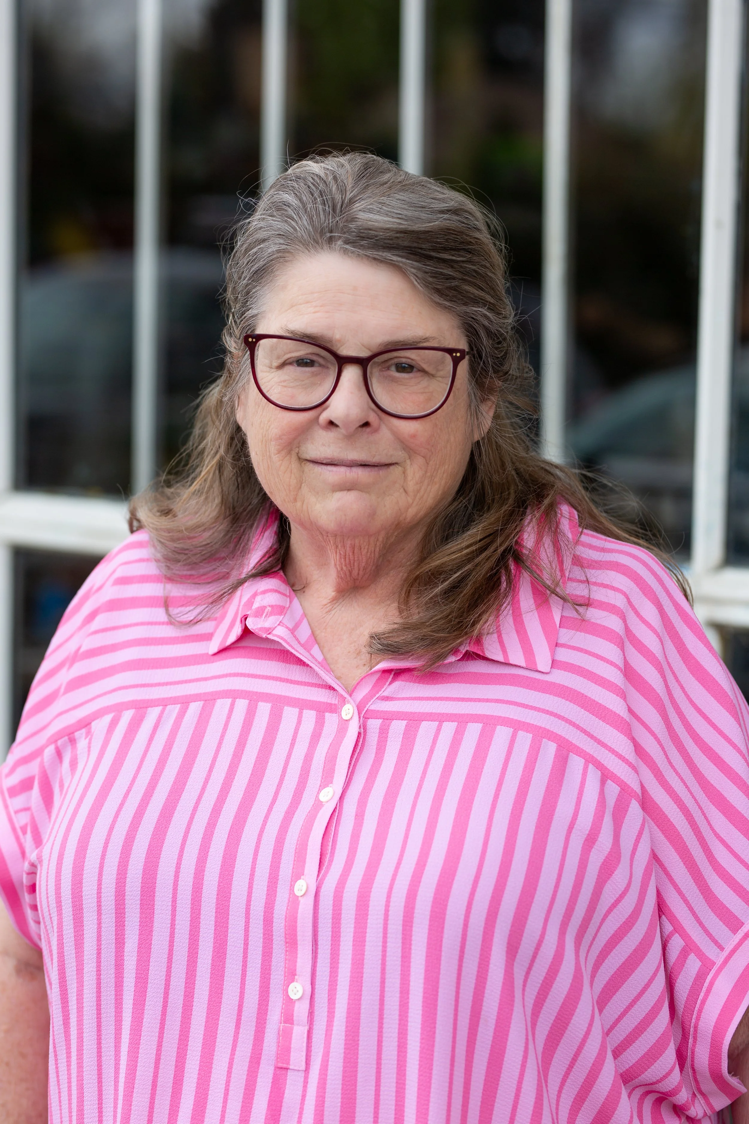 Close-up of a woman with gray hair, wearing glasses and a pink and white striped shirt, standing outdoors near a railing.