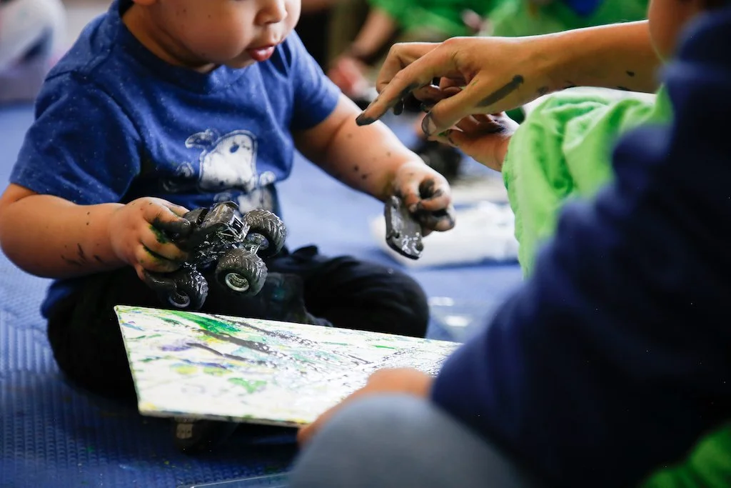 A young child holding a toy truck, with their hands covered in paint, sitting on a blue mat while an adult with paint on their hands interacts with them. The child appears focused on an artwork or project placed on the mat.