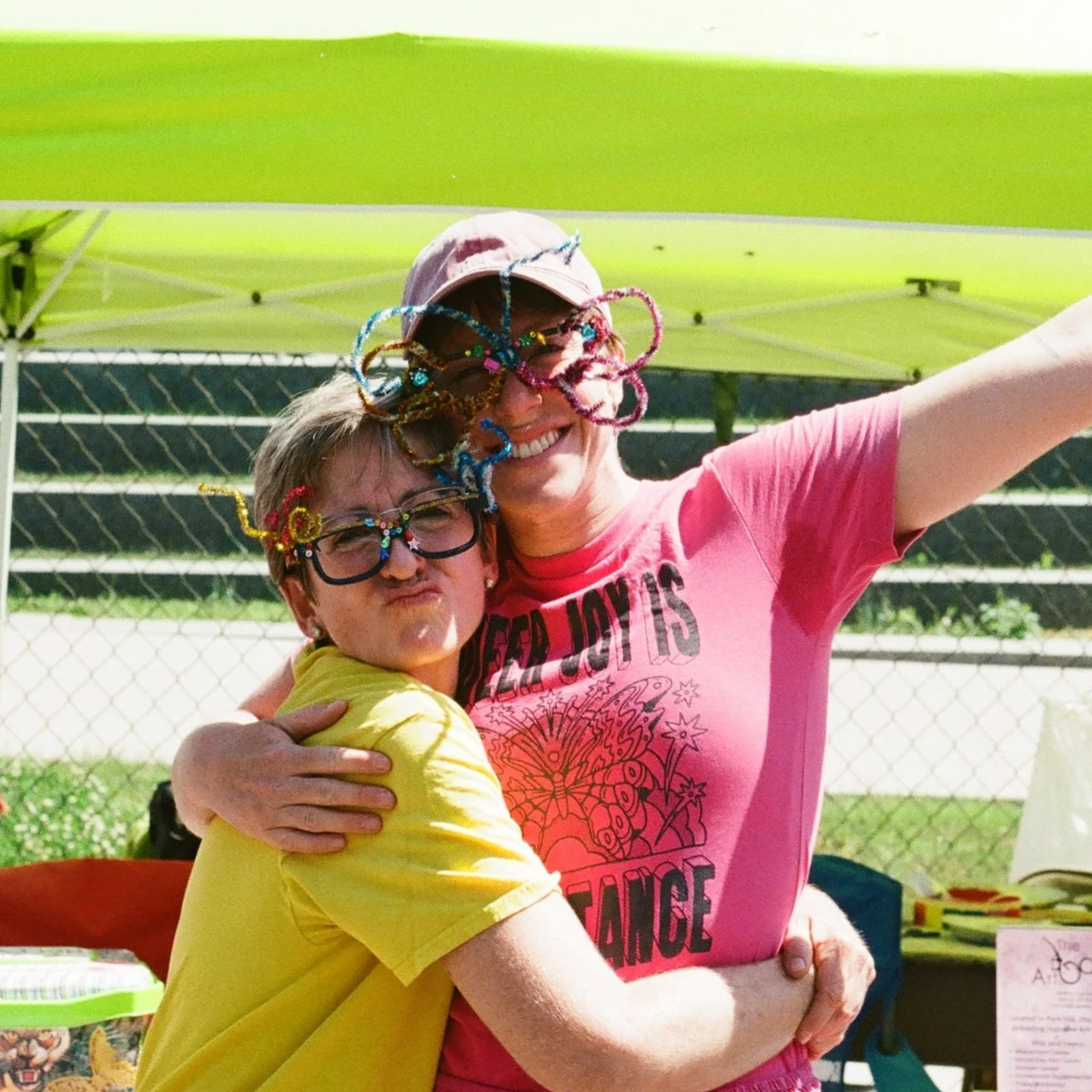 Two women hugging at an outdoor event, both wearing glasses decorated with colorful pipe cleaners and beads, smiling and celebrating.