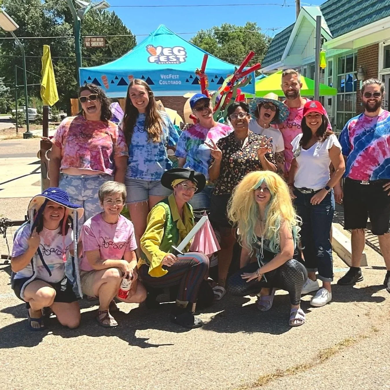 Group of people dressed in colorful, tie-dye clothing and costumes at an outdoor event. They are smiling and posing in front of a blue VegFest Colorado tent, with some wearing wigs, hats, and glasses, and a person dressed as a clown holding a sword. It is sunny with trees and houses in the background.