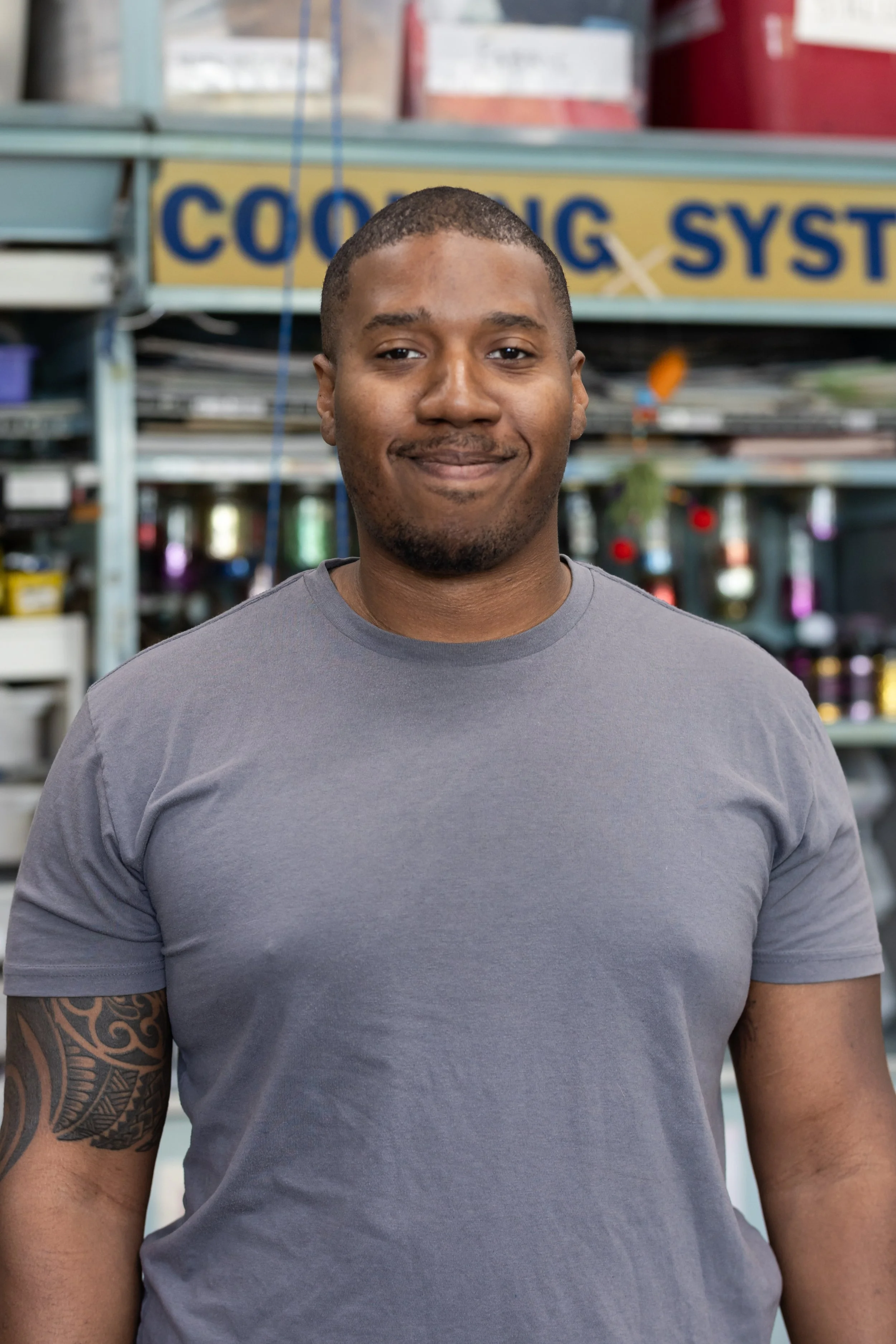 A man with a short haircut, a tattoo on his left arm, smiling slightly, wearing a gray t-shirt, standing inside a store with shelves filled with various items in the background.