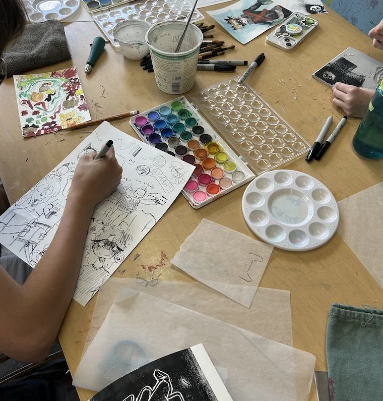 A table with watercolor paints, brushes, and paper featuring various artwork, including a colorful animal illustration and a black-and-white drawing. Hands are seen sketching and painting, surrounded by art supplies.