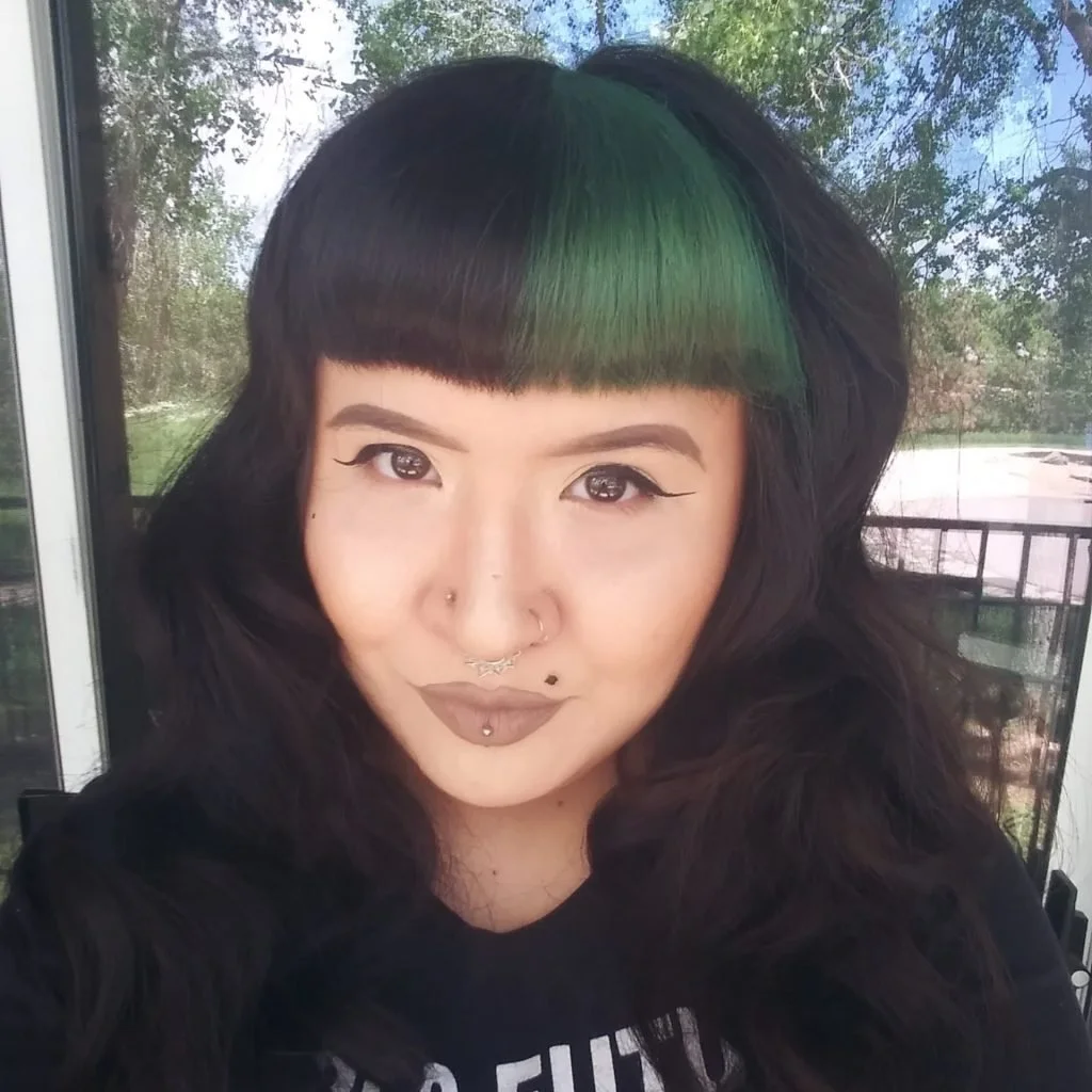 Close-up of a woman with black hair, green streaks, and a blunt fringe, smiling outdoors with trees in the background.