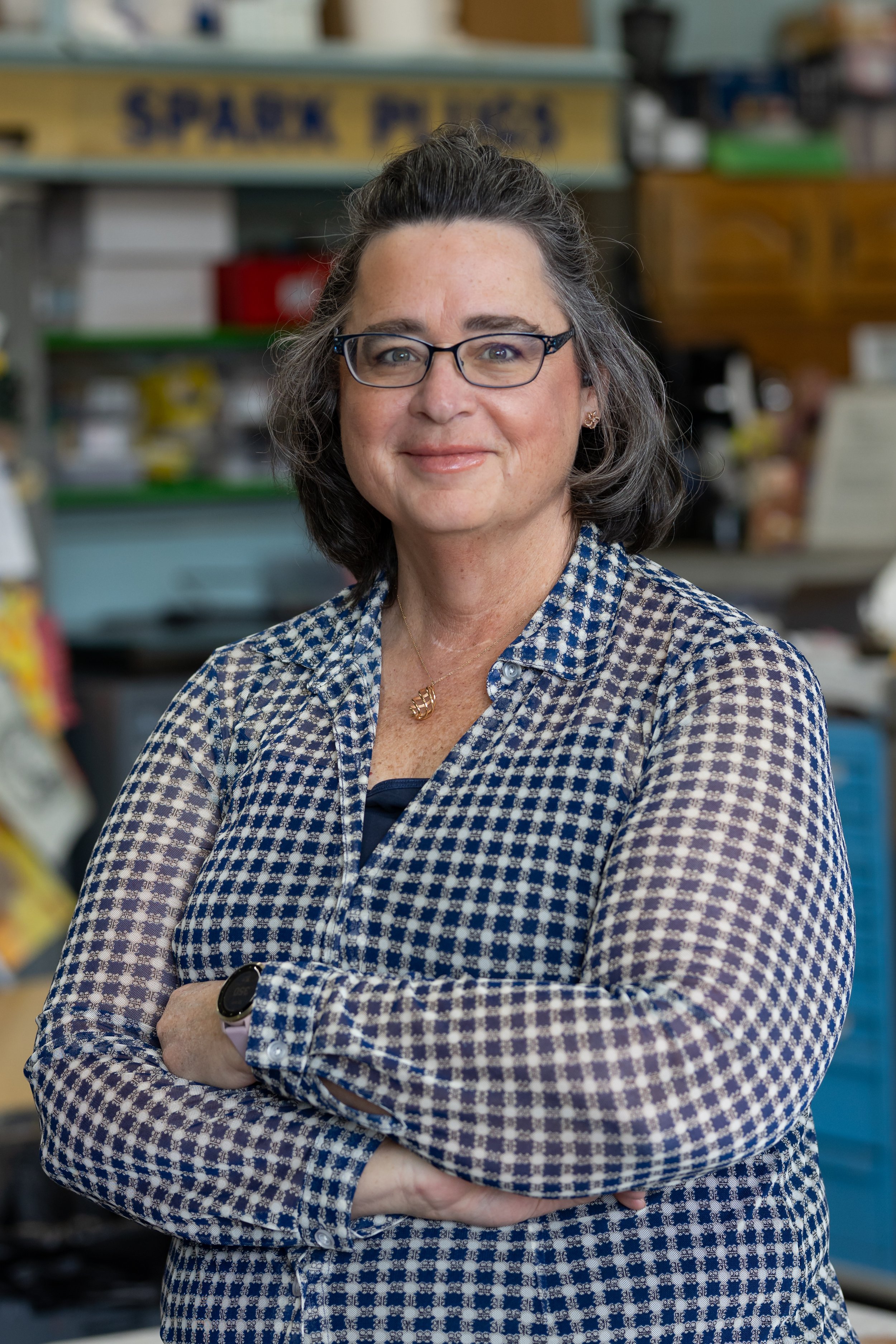 A smiling middle-aged woman with glasses, wearing a blue and white patterned shirt, standing with her arms crossed in a store aisle.