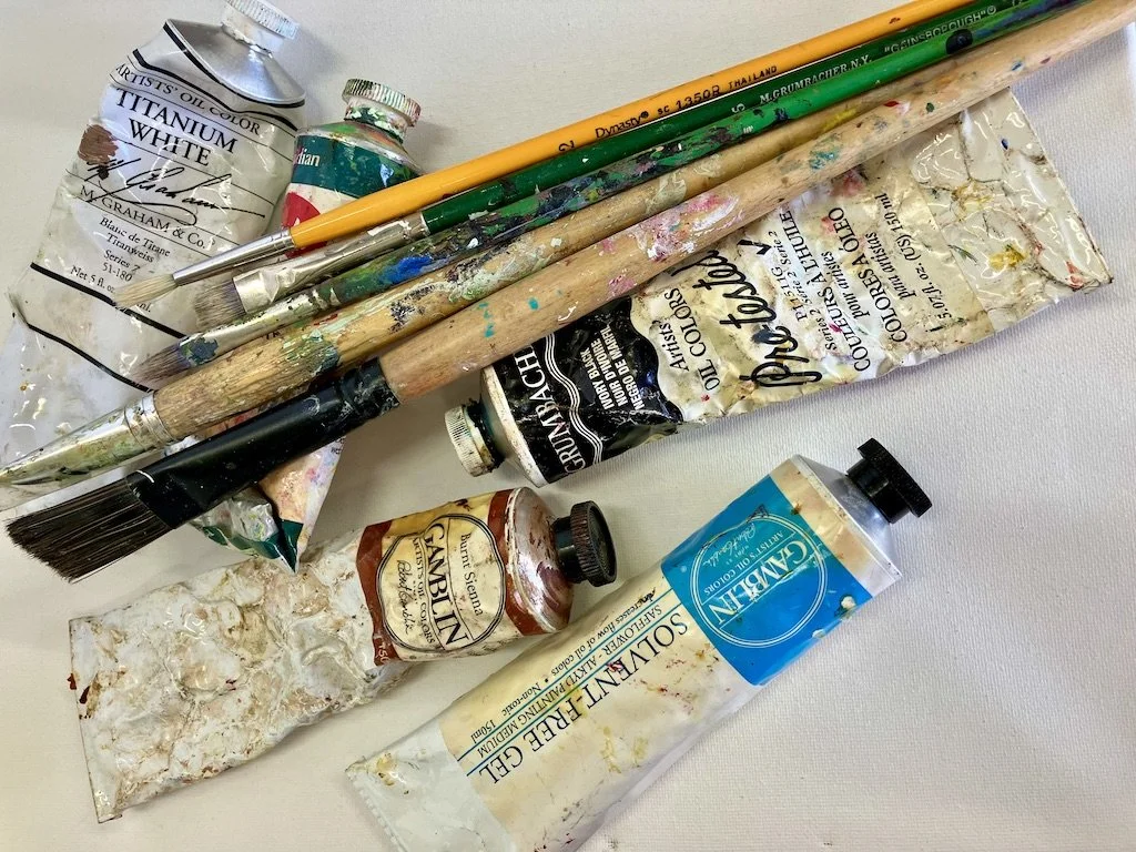 Art supplies including tubes of acrylic paint, paintbrushes, a palette knife, and a crumpled plastic palette with paint on it.