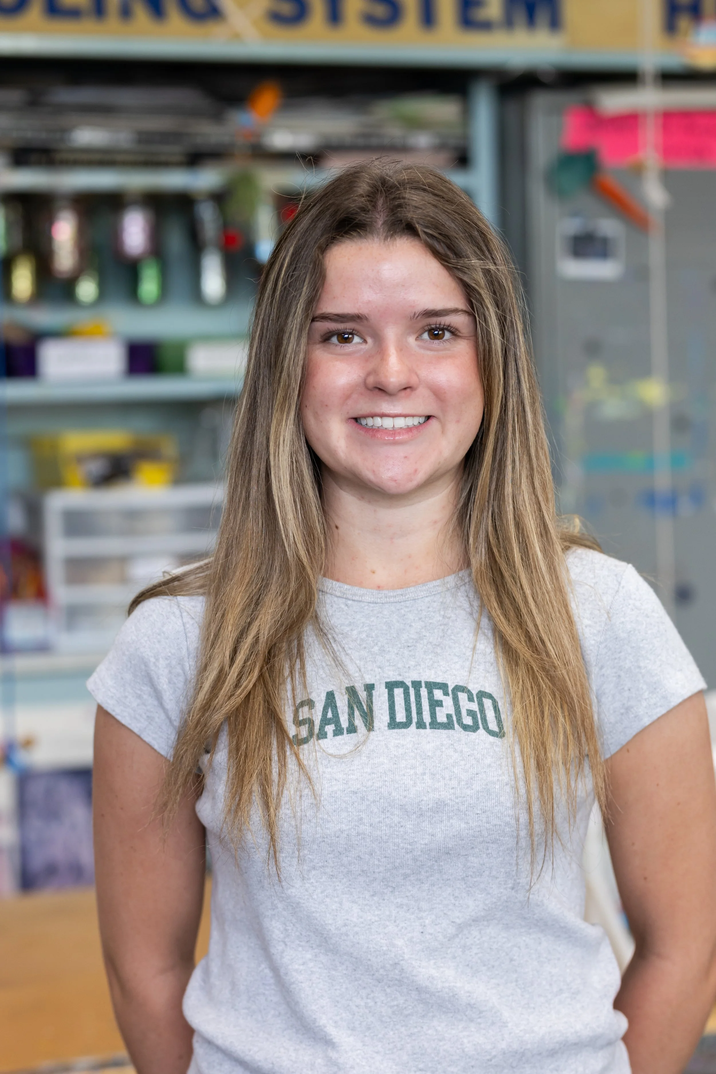 A young woman with long brown hair and a light gray T-shirt with green text reading 'SAN DIEGO' on it, standing indoors in front of a blurred background of shelves filled with bottles and various items.