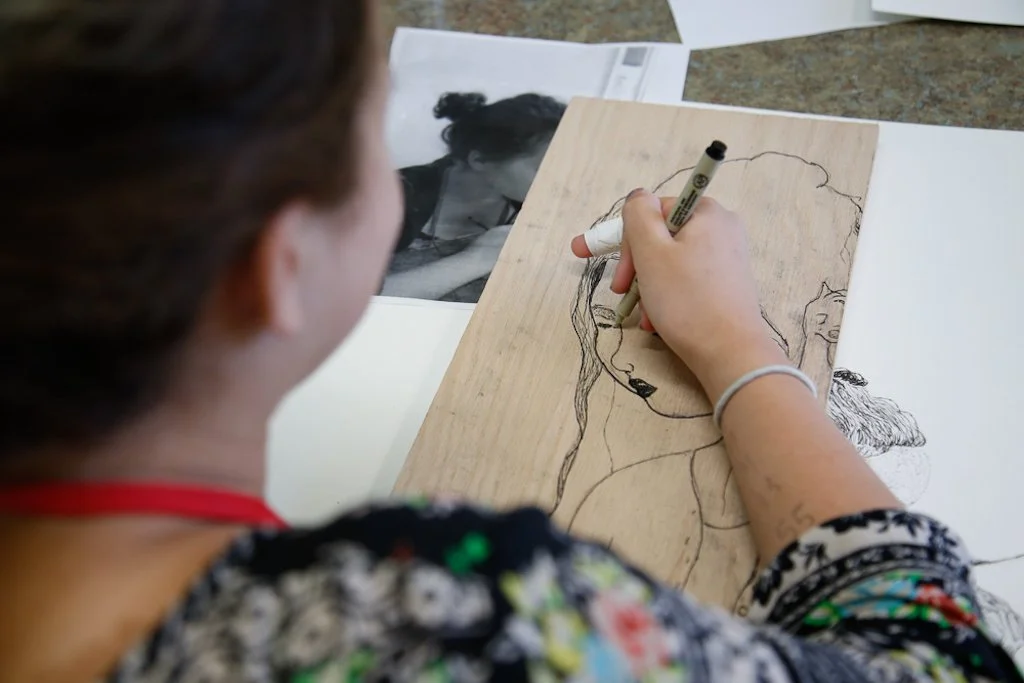 A person is drawing a portrait on a wooden board with a black marker, with a printed photo of a woman beside them.
