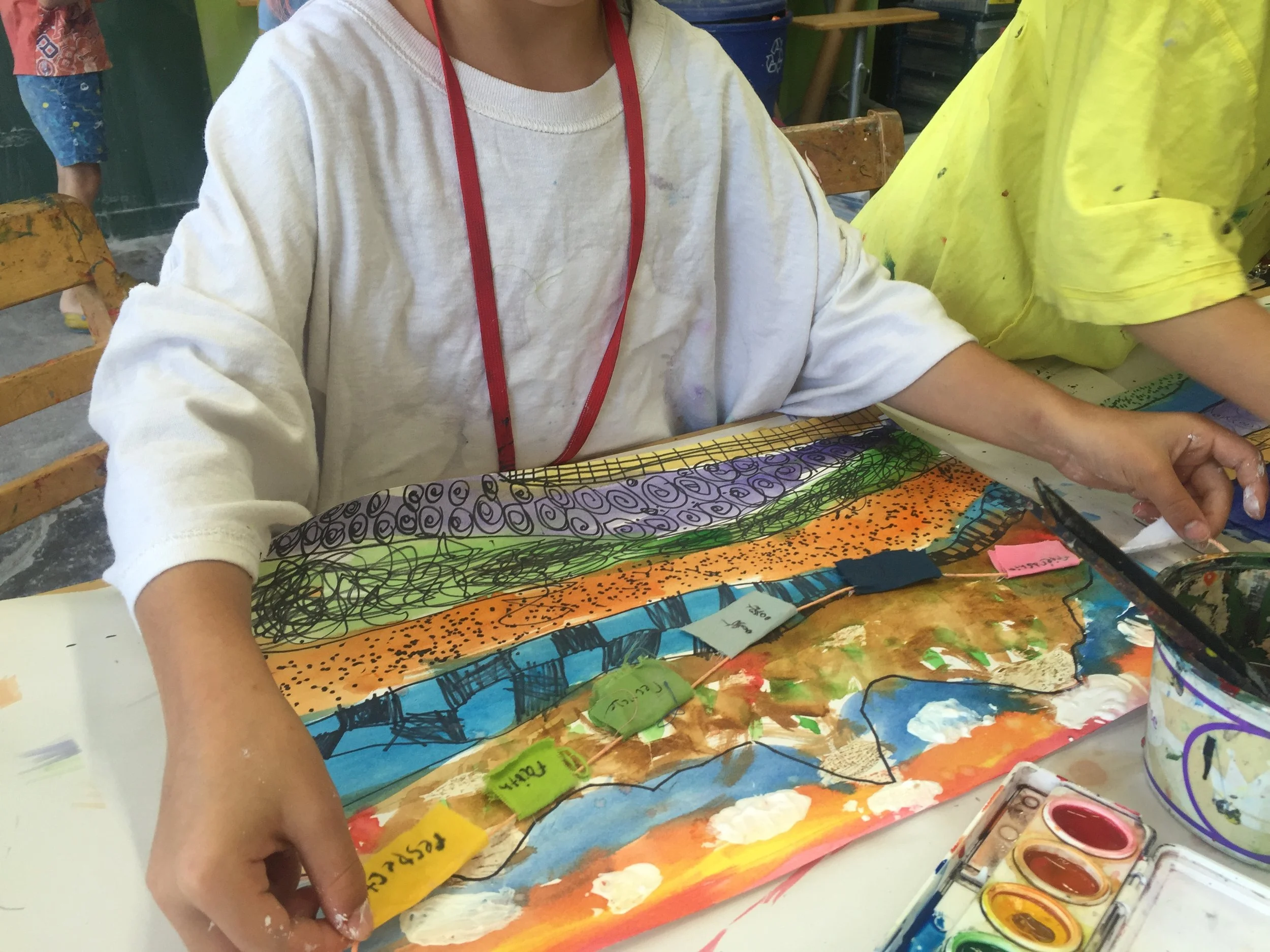 A person in a white long-sleeve shirt with a red lanyard sitting at a table, engaged in painting a colorful artwork with paint palettes visible in the foreground. The background shows other people, some wearing bright yellow and blue clothing.