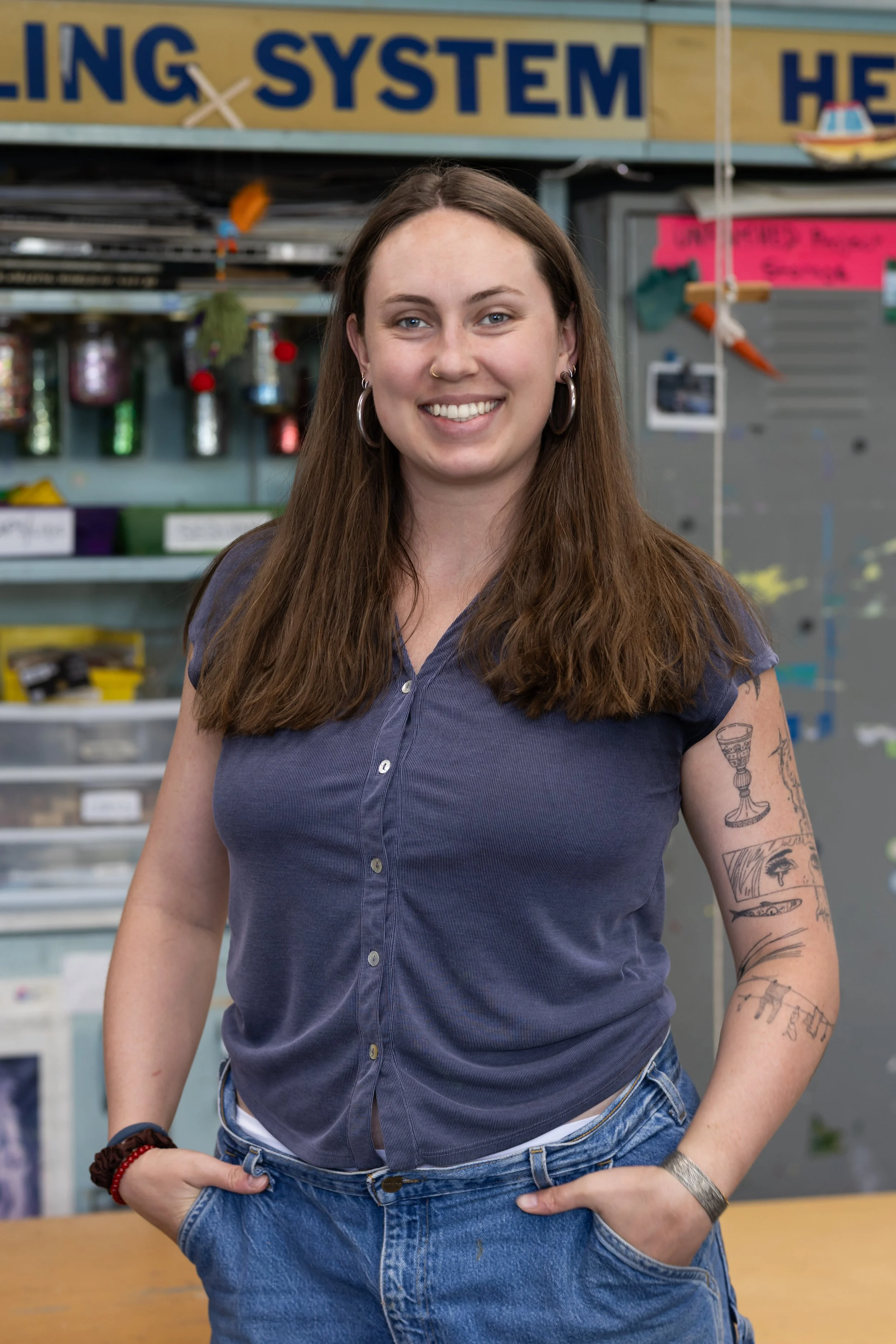 Young woman with long brown hair smiling, wearing a blue shirt and jeans, has tattoos on her arm, standing in front of a cluttered workshop or hardware store.