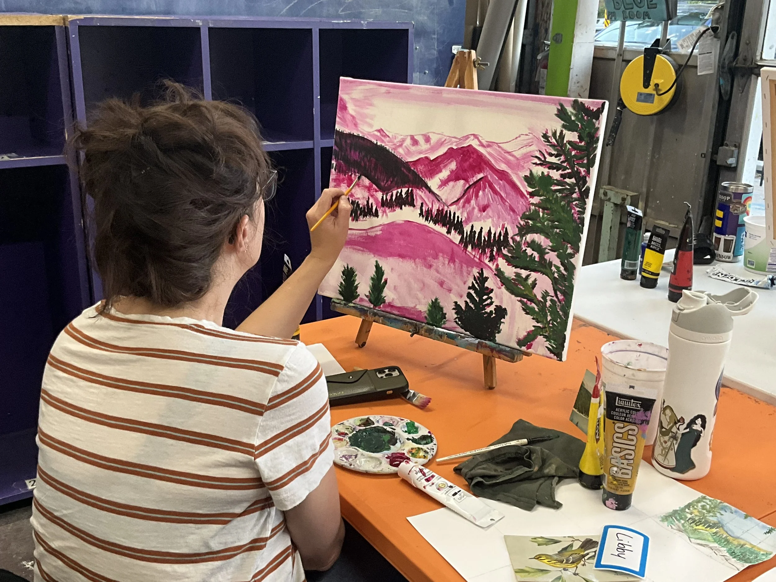 A person with brown hair wearing a white and orange striped shirt painting a landscape scene with mountains, trees, and pink sky on a canvas at an art station.