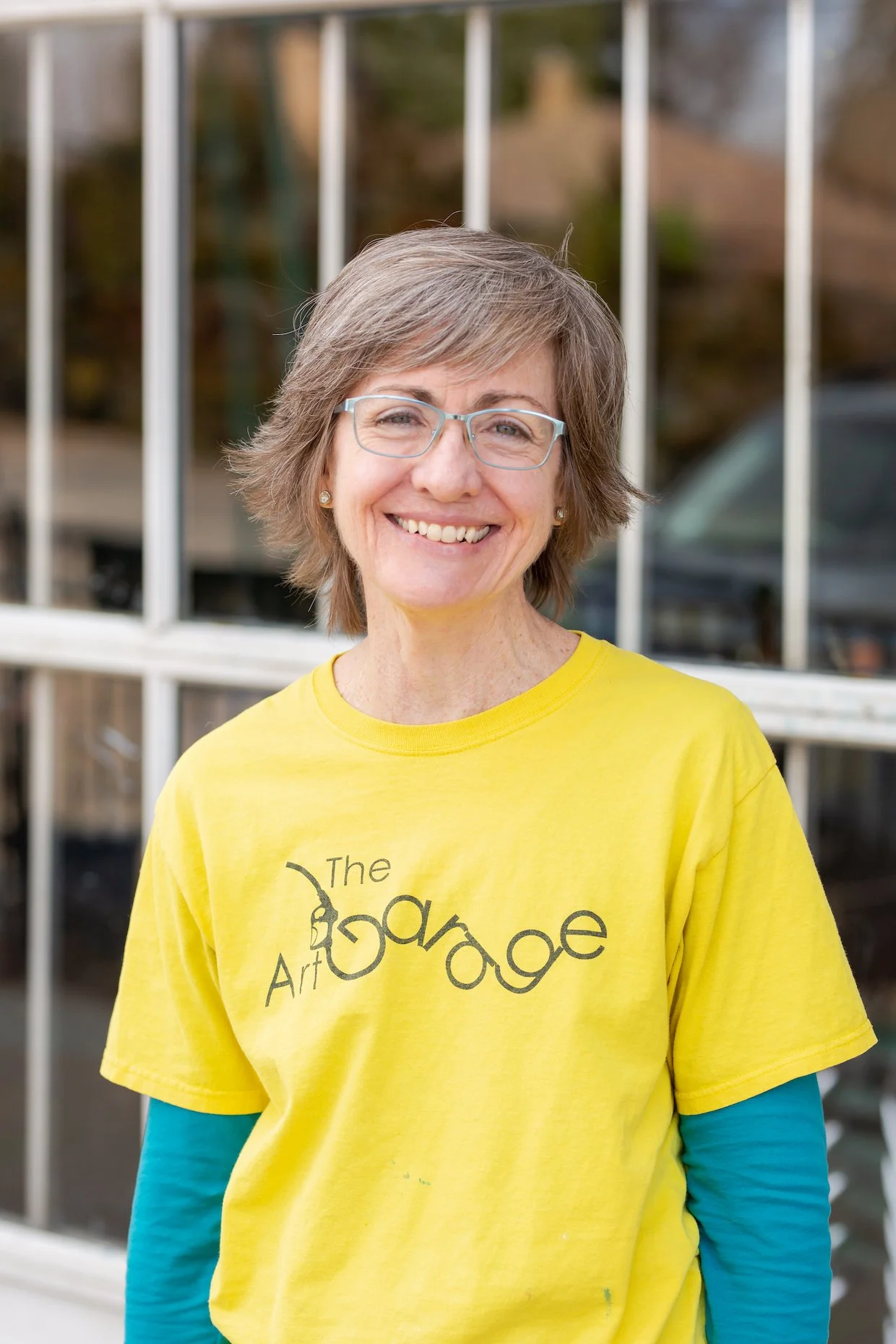 A smiling woman with short gray hair and glasses, wearing a yellow T-shirt and a blue long-sleeve shirt underneath, standing outdoors in front of a building with white window frames.