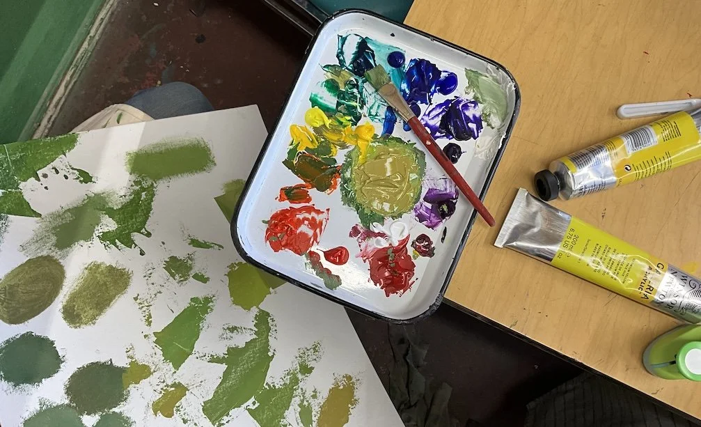 Paint palette with various colors and a paintbrush, next to a sheet of paper with green paint strokes, on a wooden table.