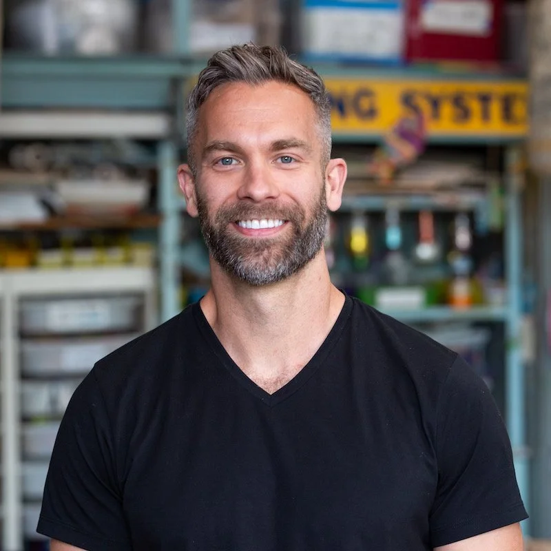 A smiling man with gray hair and a beard, wearing a black V-neck T-shirt, standing in a workshop or industrial setting with shelves of tools and equipment in the background.