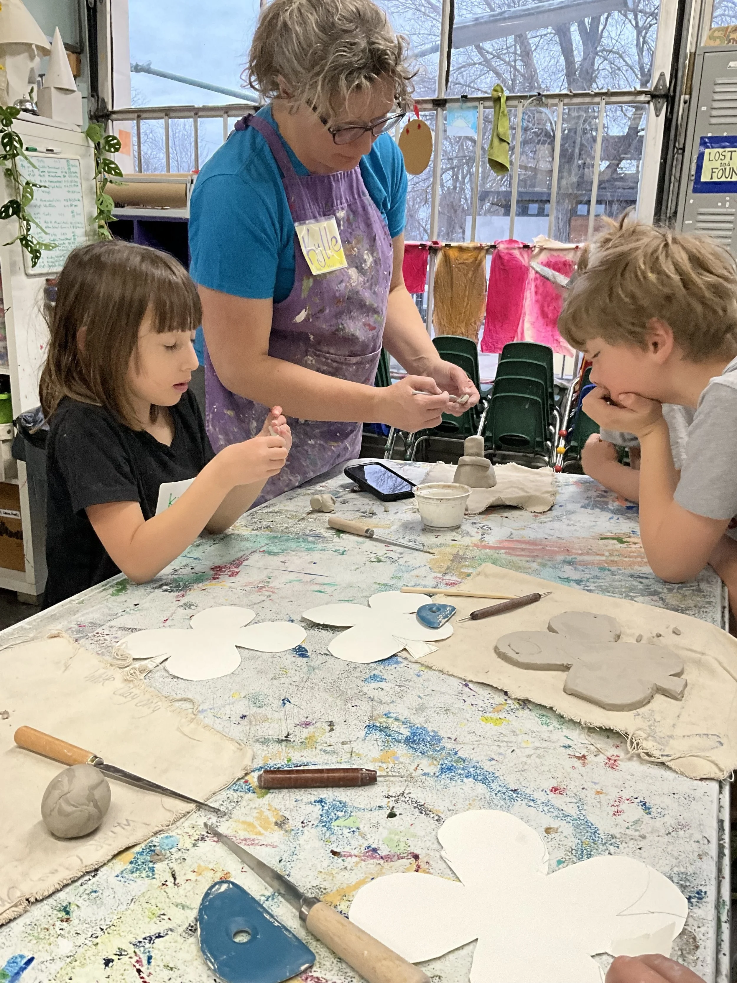 An art classroom with two children and an instructor working on clay art projects, various tools, and paper cutouts on the table.