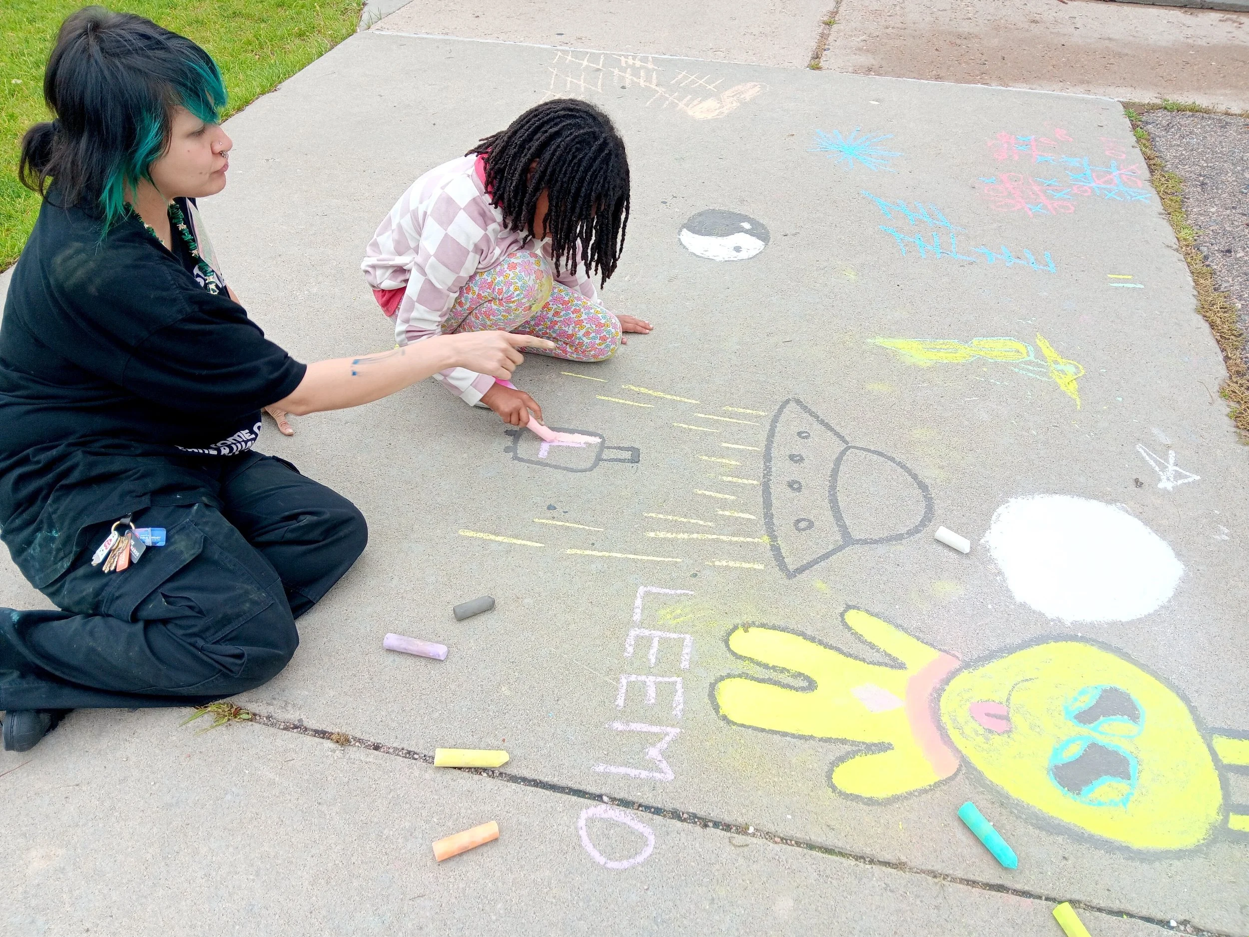 Two girls are sitting on a sidewalk drawing with chalk. One girl has black hair with teal highlights, and the other girl has dark dreadlocks and is wearing floral pants. They are creating colorful chalk drawings on the pavement which include various shapes, patterns, and a rocket ship, with chalk pieces scattered nearby.