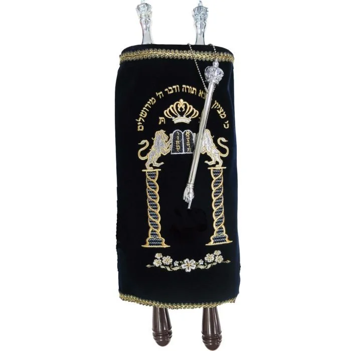 A ceremonial Torah scroll with an ornate velvet cover embroidered with Hebrew text, golden and silver decorative elements, two silver torches, and a silver scepter, all on a wooden platform.