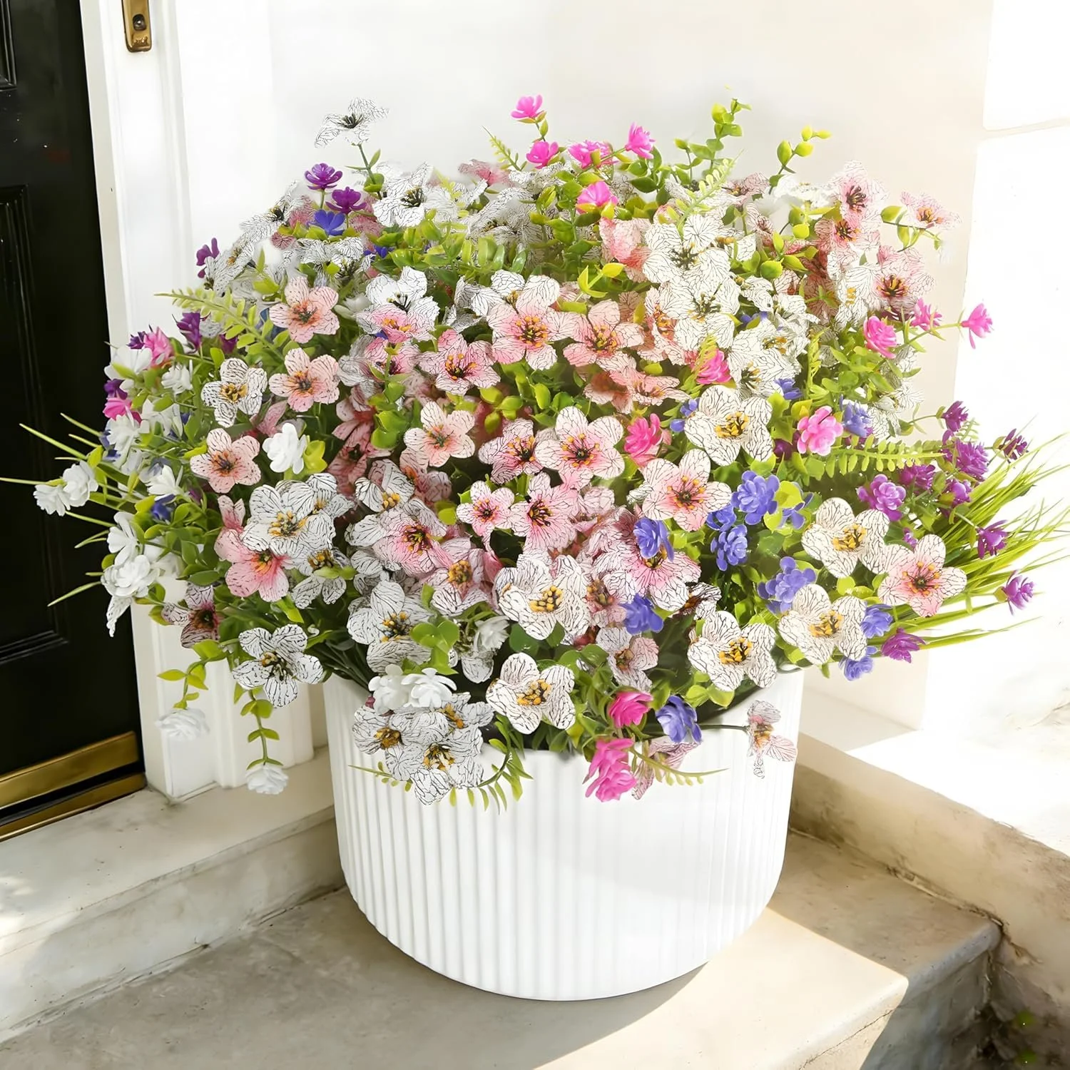 A large white pot filled with colorful artificial flowers including pink, purple, white, and blue flowers placed near a black door and white marble steps.