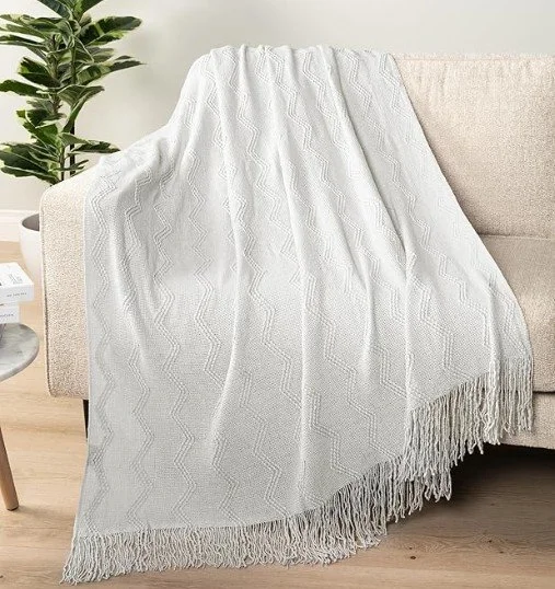A white textured throw blanket with fringes draped over the arm of a beige sofa in a living room.