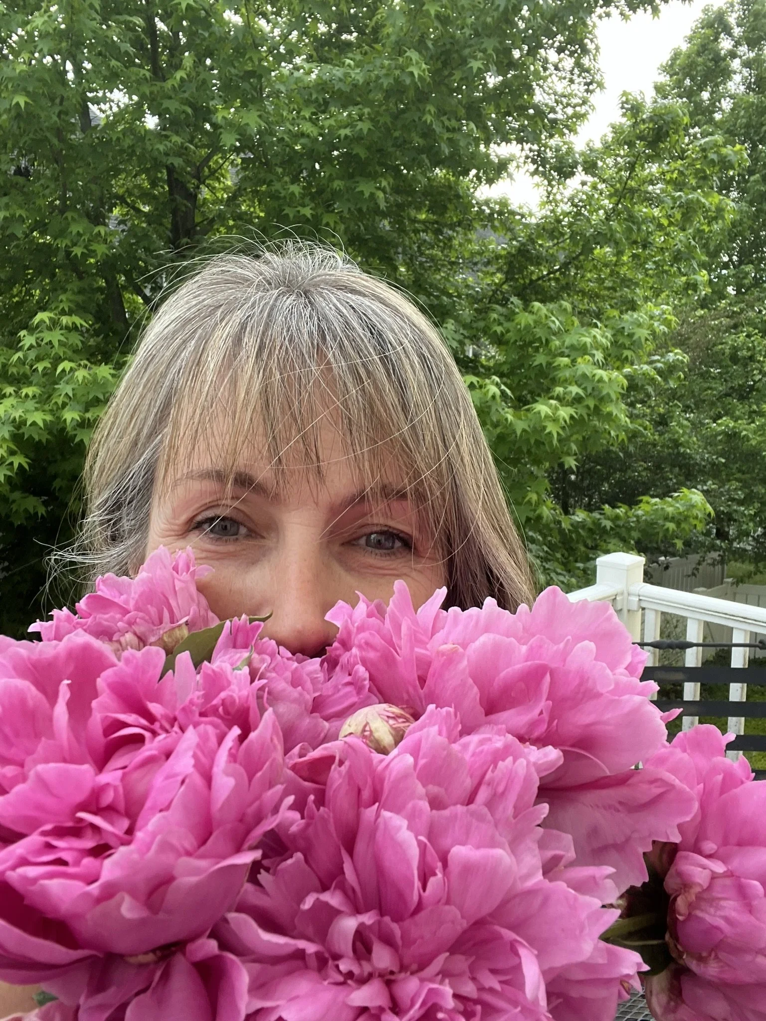 Partially visible woman with gray hair holding a large pink peony flower in front of her face, outdoors in a garden with green trees and a white fence in the background.