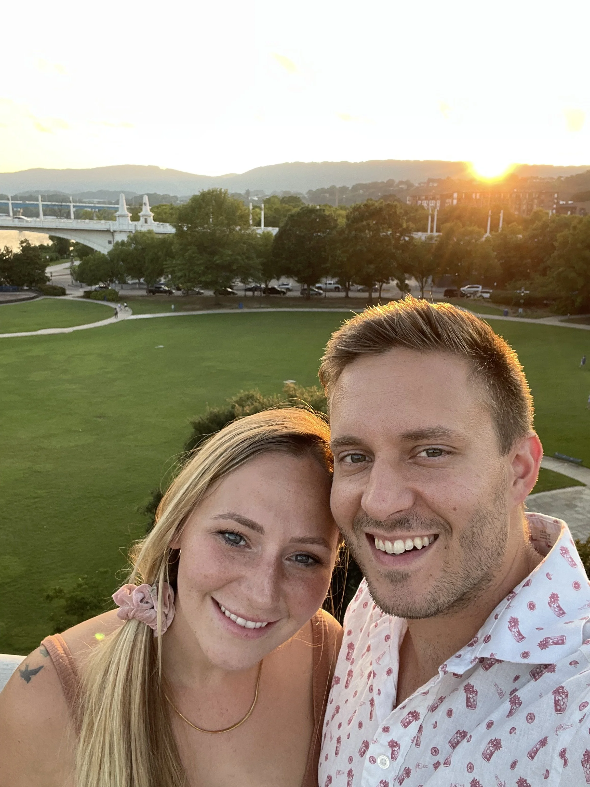 Close-up of a smiling young woman with blonde hair and a man with short hair, smiling, outdoors during sunset with green grass, trees, a bridge, hills, and buildings in the background.