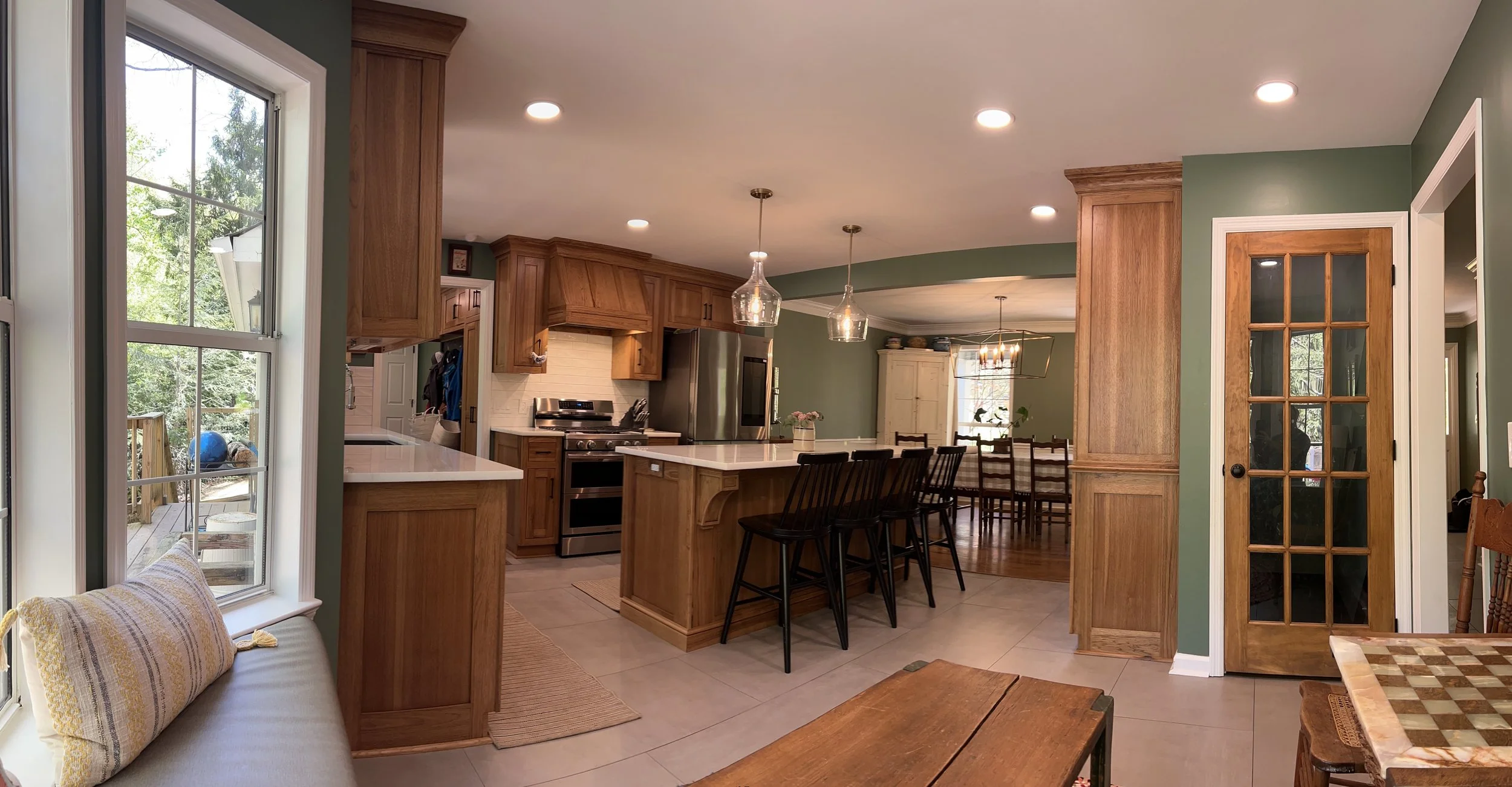 Kitchen with wooden cabinets, island, stainless steel stove, and dining area with wooden table and chairs, green walls, and large windows showing outdoor trees.