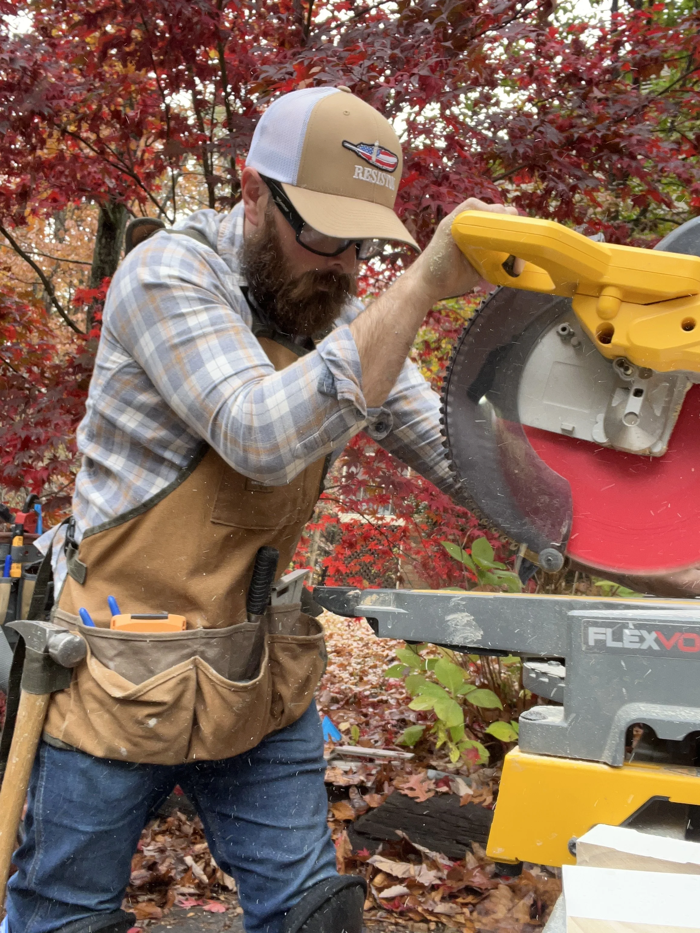 A person wearing a beige and white trucker hat with a patch that says 'RESIST' and features a patriotic design, glasses, a plaid shirt, and a brown work apron with tools, using a yellow power saw to cut wood outdoors with red and green autumn foliage in the background.