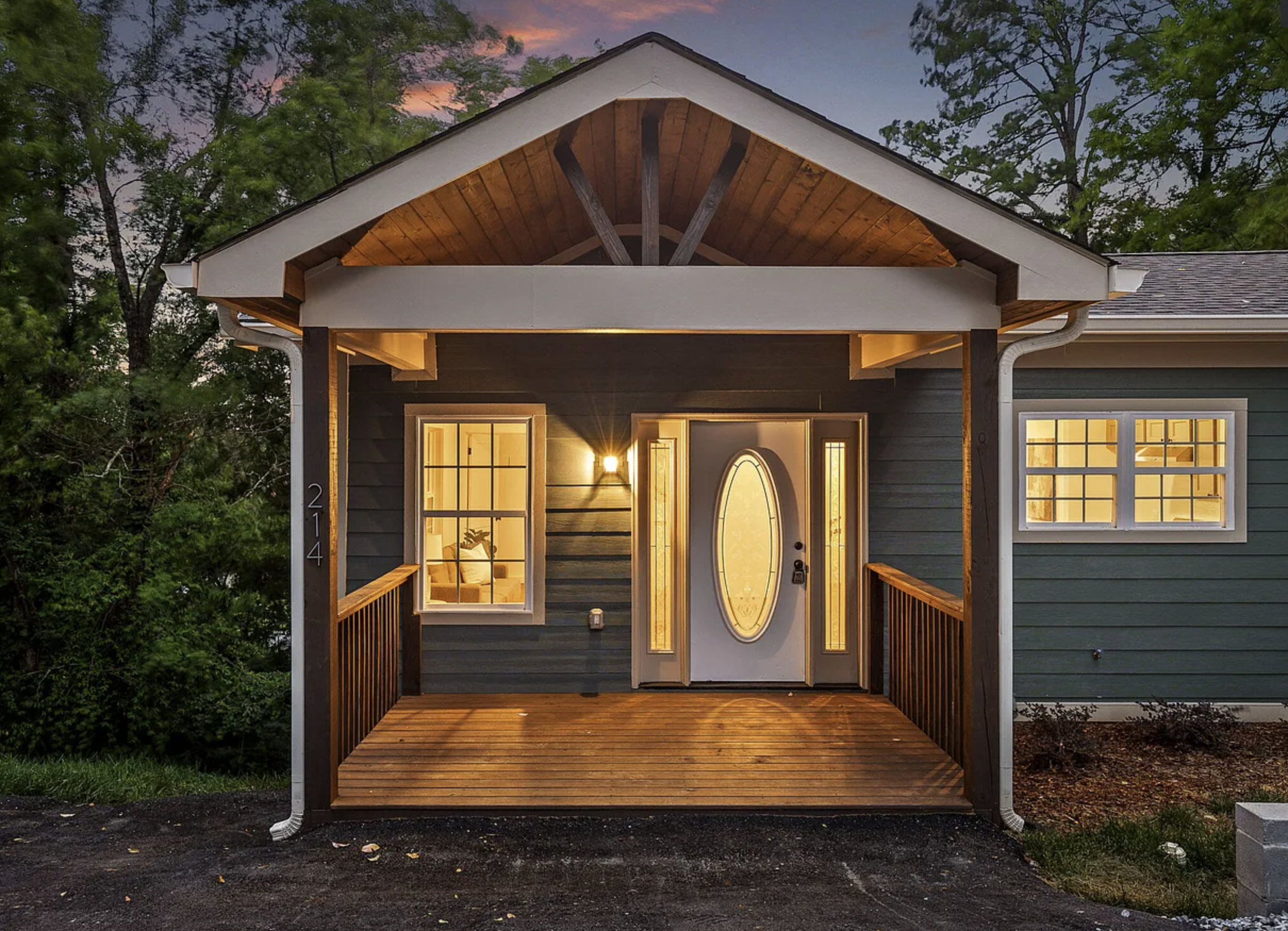 Front view of a house with a covered porch, illuminated from inside, with large windows and a gabled roof, surrounded by trees during dusk.