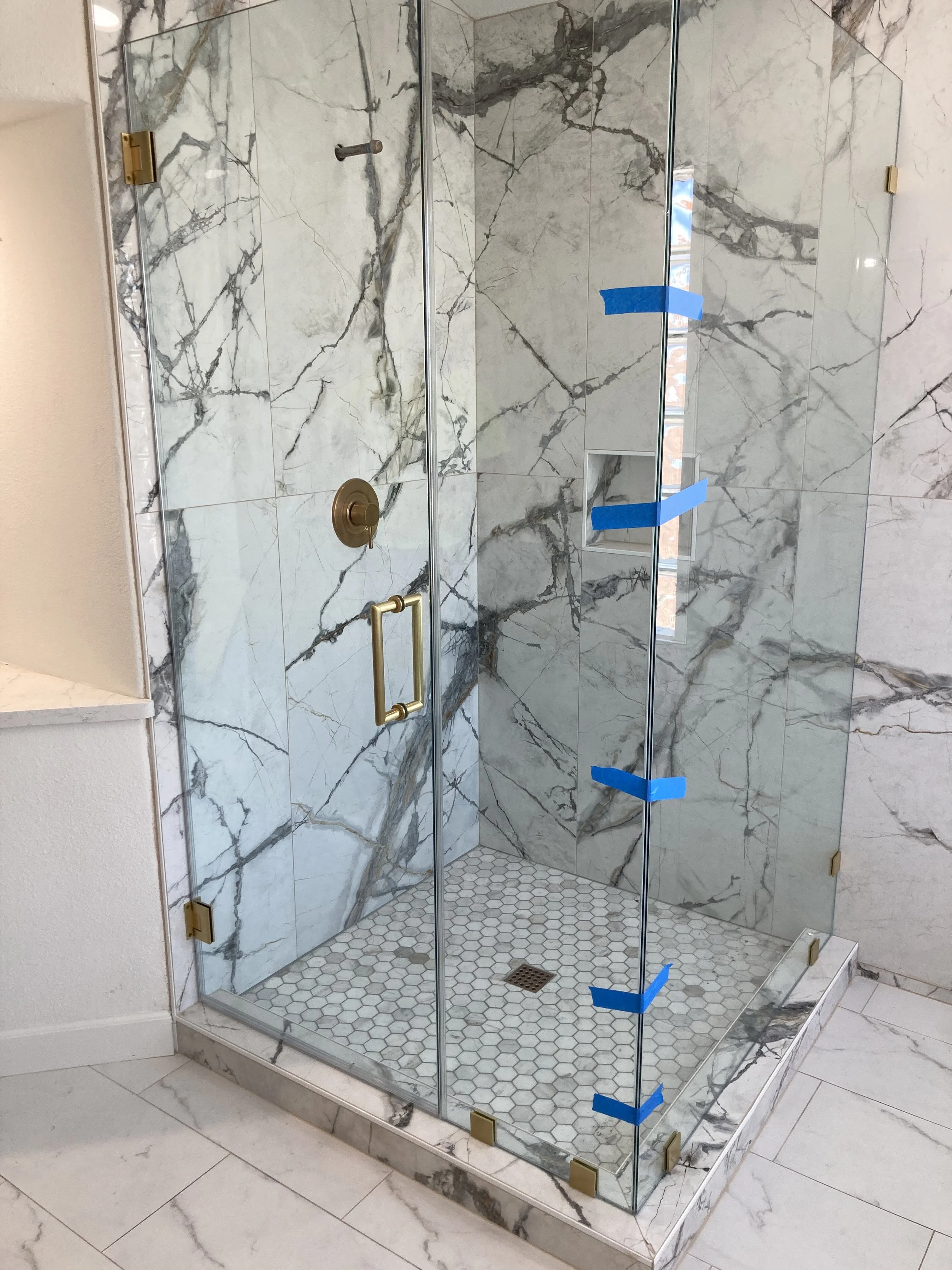 A glass shower enclosure with marble walls and a small hexagonal tiled shower floor. The enclosure has a gold handle, gold hinges, and a built-in niche in the wall. Blue painter's tape is applied to parts of the glass, likely for protection or marking during installation.
