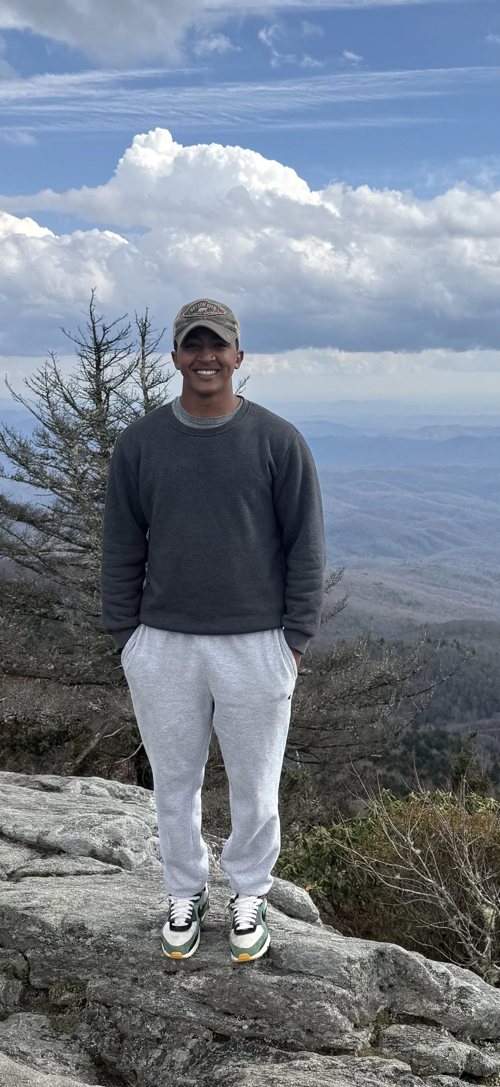 Young man standing on a rocky outcrop with a mountainous landscape and blue sky with clouds in the background.