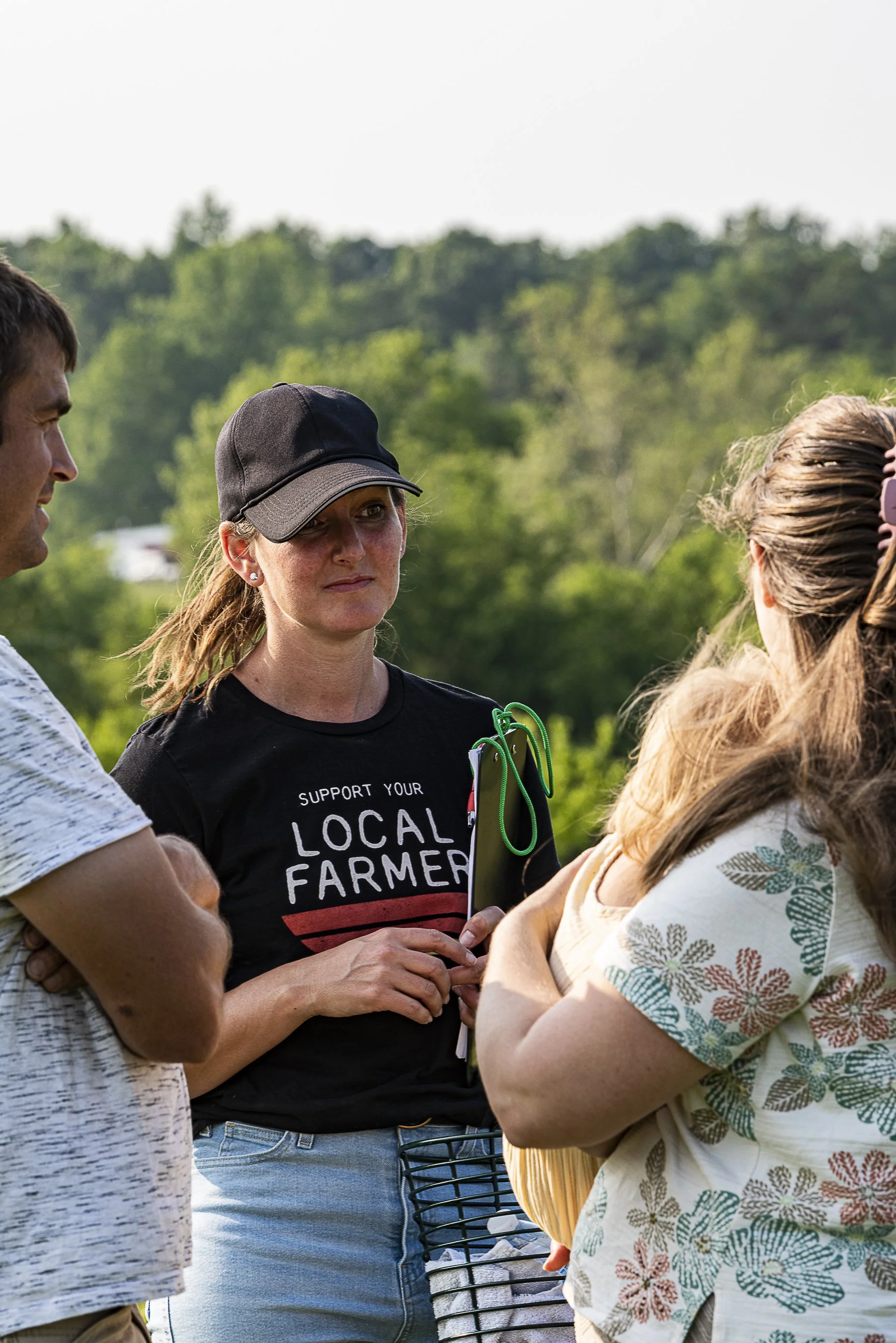 Four people are gathered outdoors having a conversation, with trees in the background. One woman wears a black cap and a T-shirt that says 'Support Your Local Farmer,' while another woman wears a floral-patterned shirt. The man on the left is partially visible.