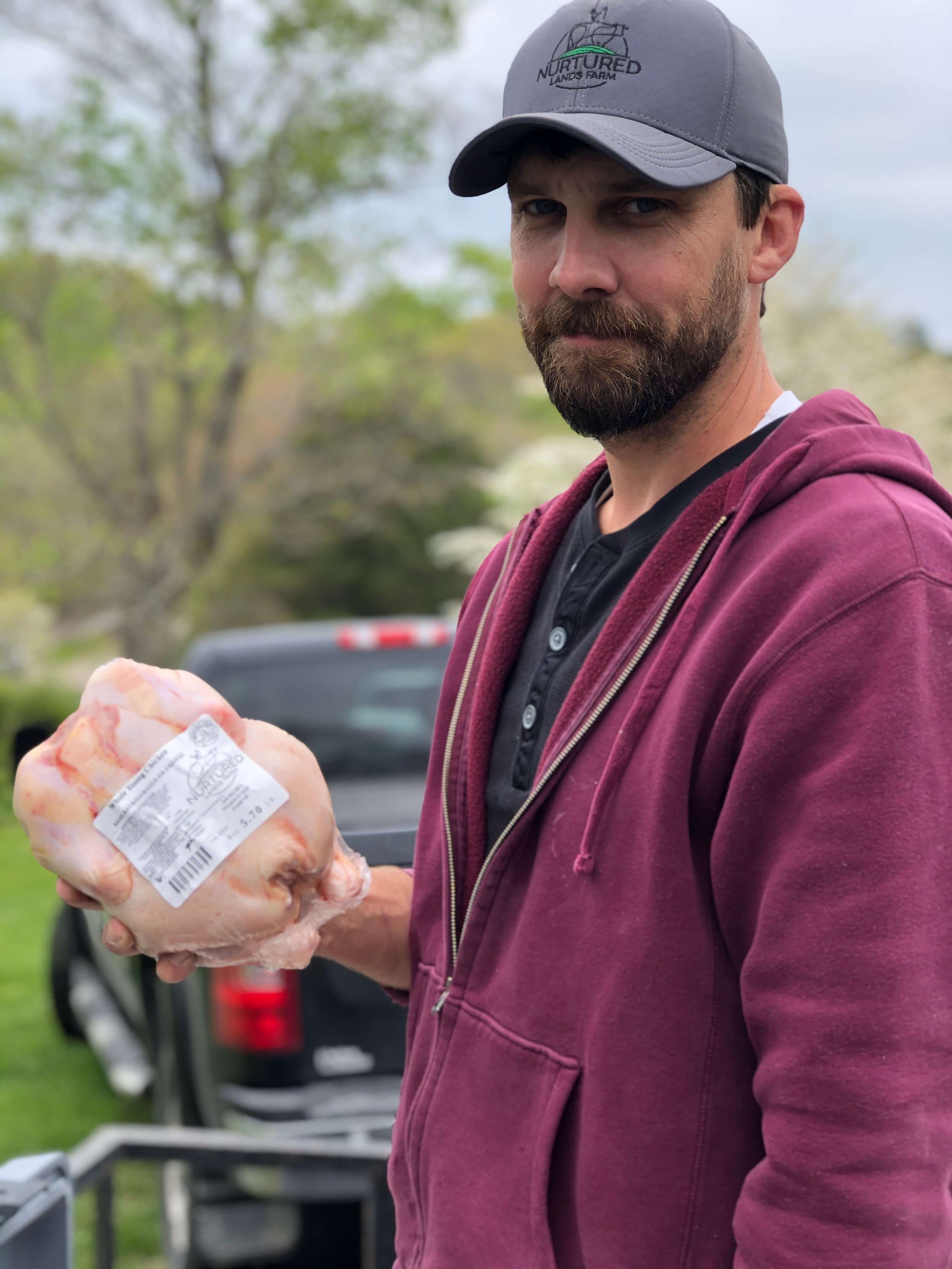 A man with a beard wearing a gray cap and a maroon hoodie holding a raw chicken with a price tag, outdoors with a blurred background of trees and a car.