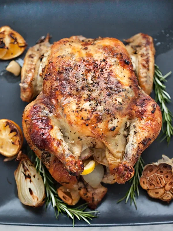 Whole roasted chicken garnished with rosemary, lemon, garlic, and grilled vegetables on a black platter.